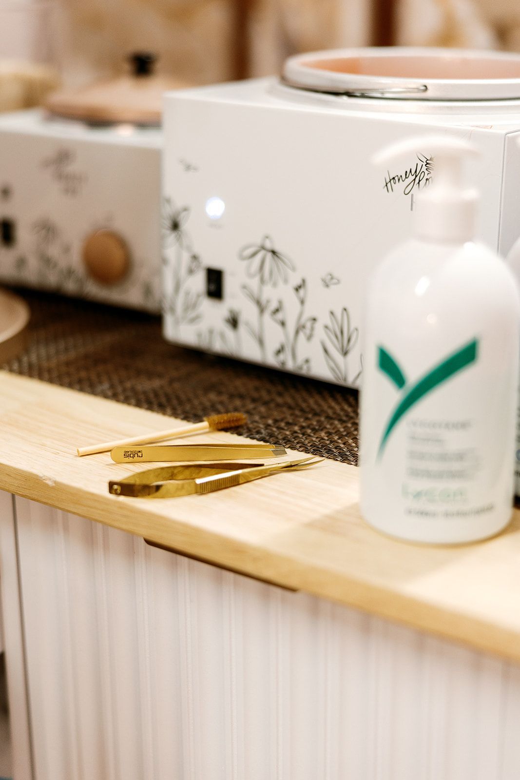 White lotion bottle beside gold tweezers on a wooden spa counter with a floral tissue box in the background