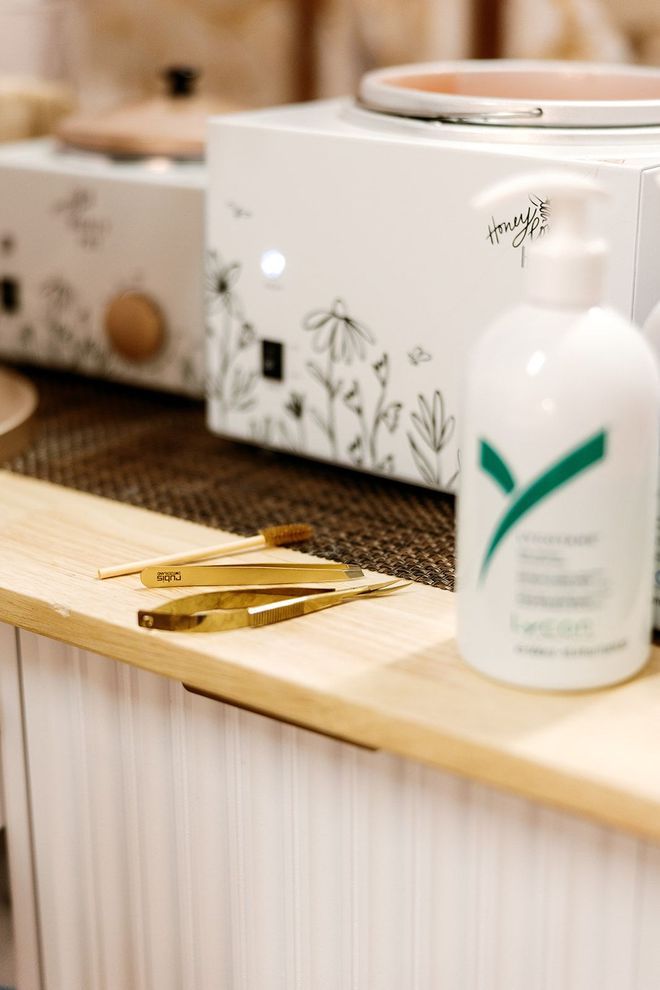 White lotion bottle beside gold tweezers on a wooden spa counter with a floral tissue box in the background