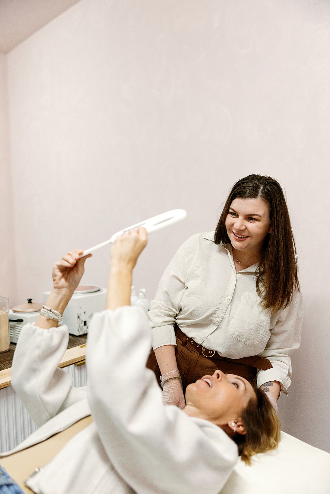 Woman lying on a treatment bed smiles while holding up a mirror toward a standing woman in a white top.