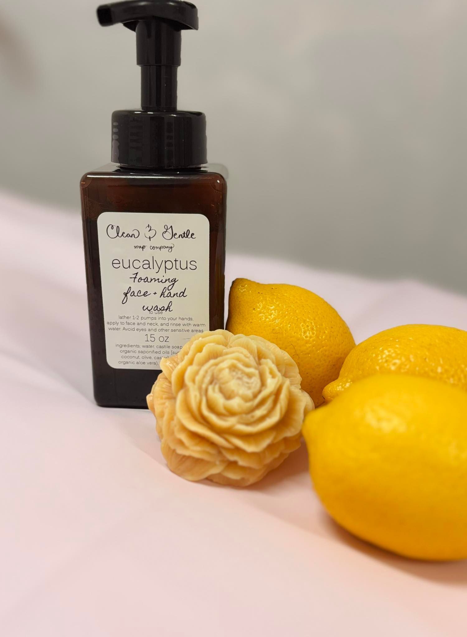 A brown soap dispenser next to lemons and a carved soap flower on a pink surface.