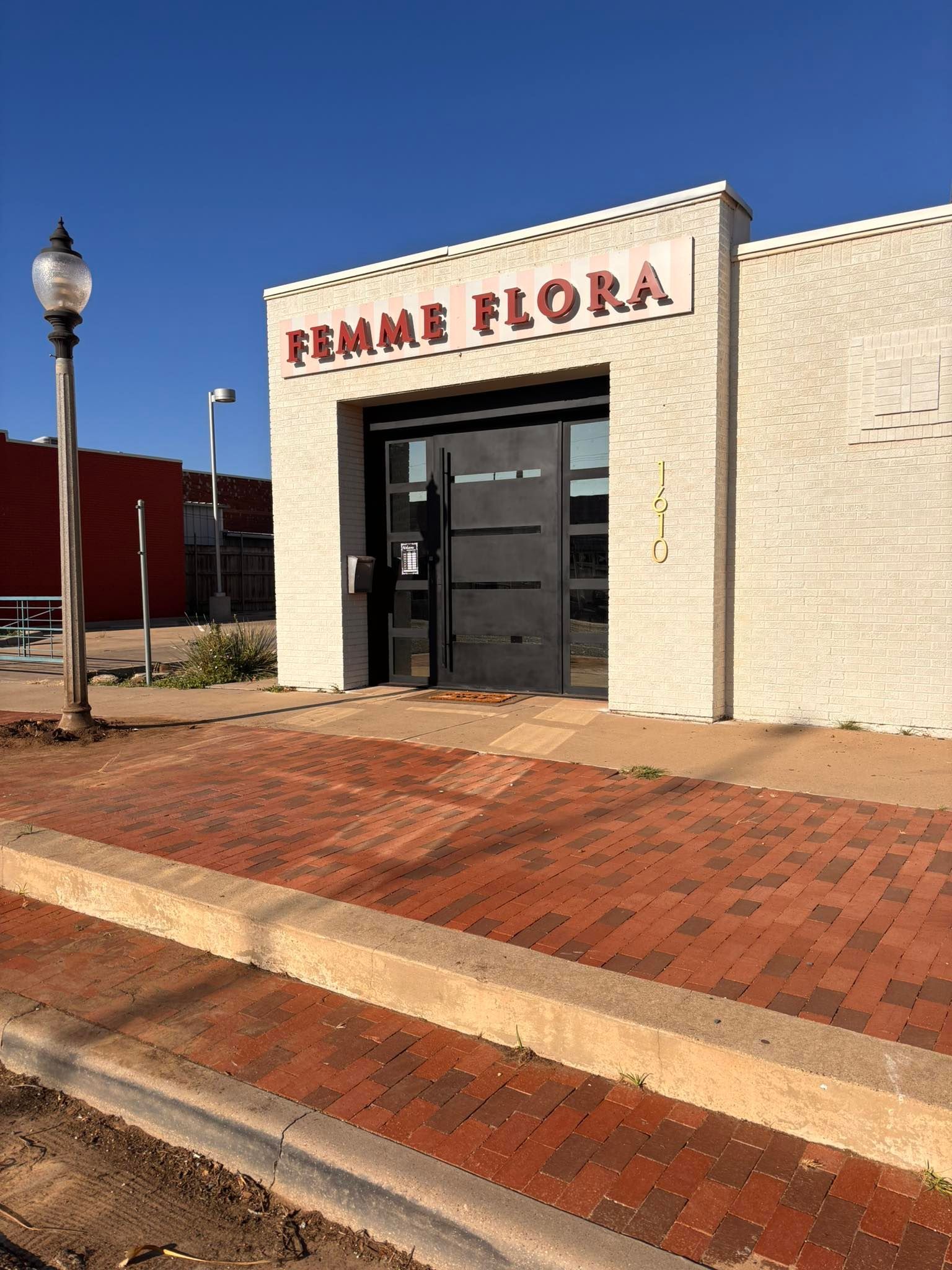 Brick storefront with red “Femme Flora” sign, black door, and brick sidewalk under a clear blue sky