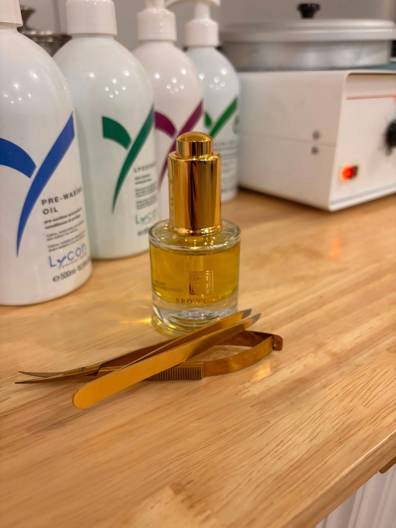 Bottle of oil, gold tweezers, and skincare products on a wooden counter in a salon.