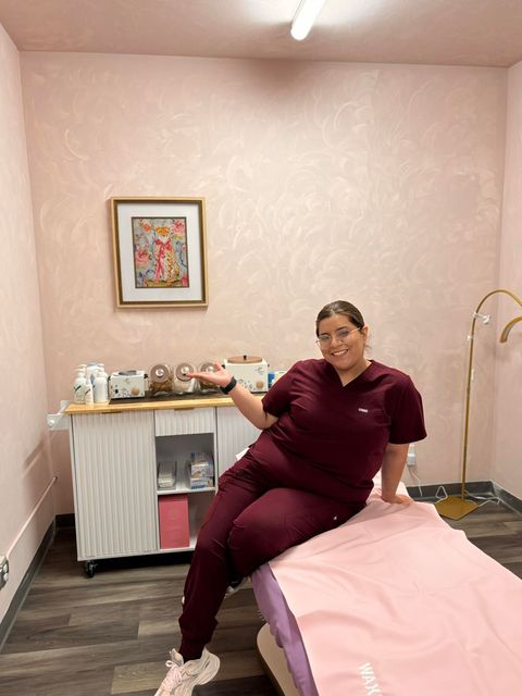 Woman in burgundy scrubs sits on a pink-covered spa bed, pointing, in a pink-walled room.