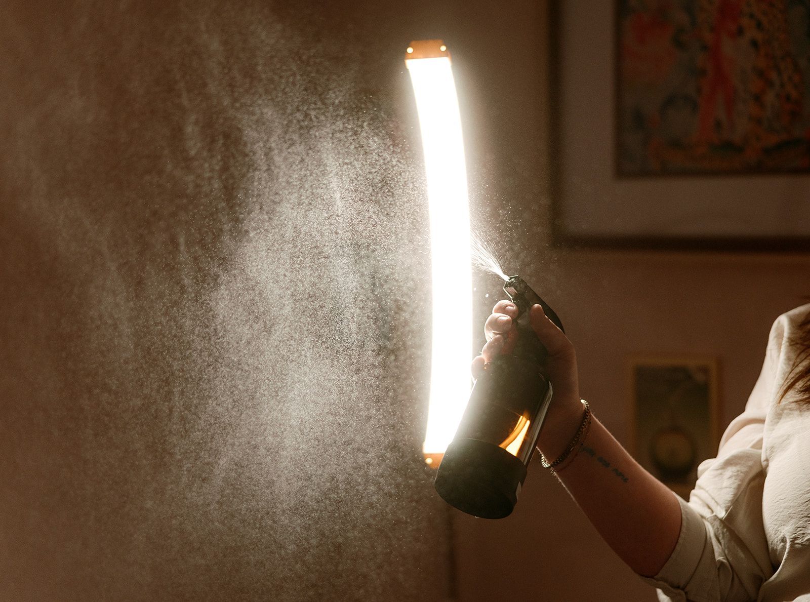 Person spraying mist from a black bottle beside a bright vertical light panel in a room