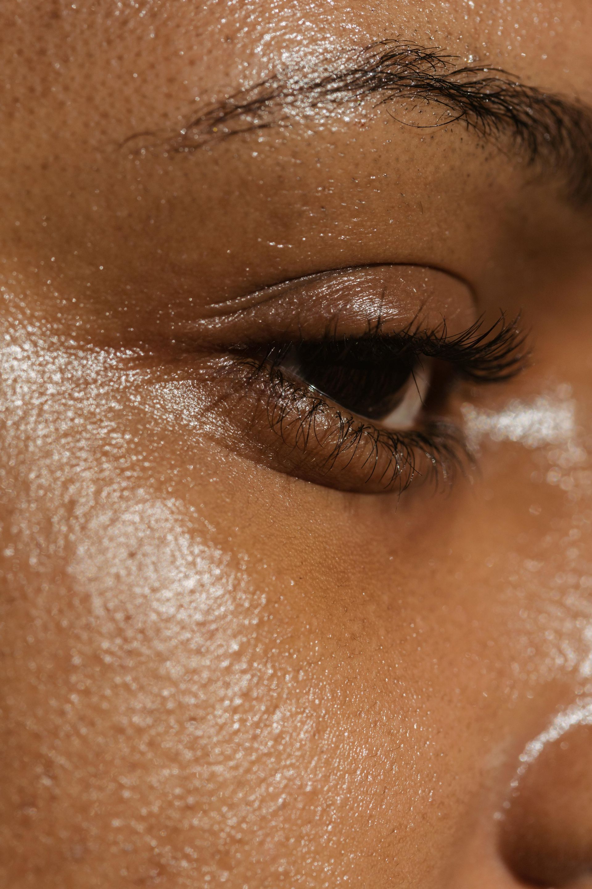 Close-up of a person's eye and face. Skin is shiny with sweat. Dark eye with black mascara.