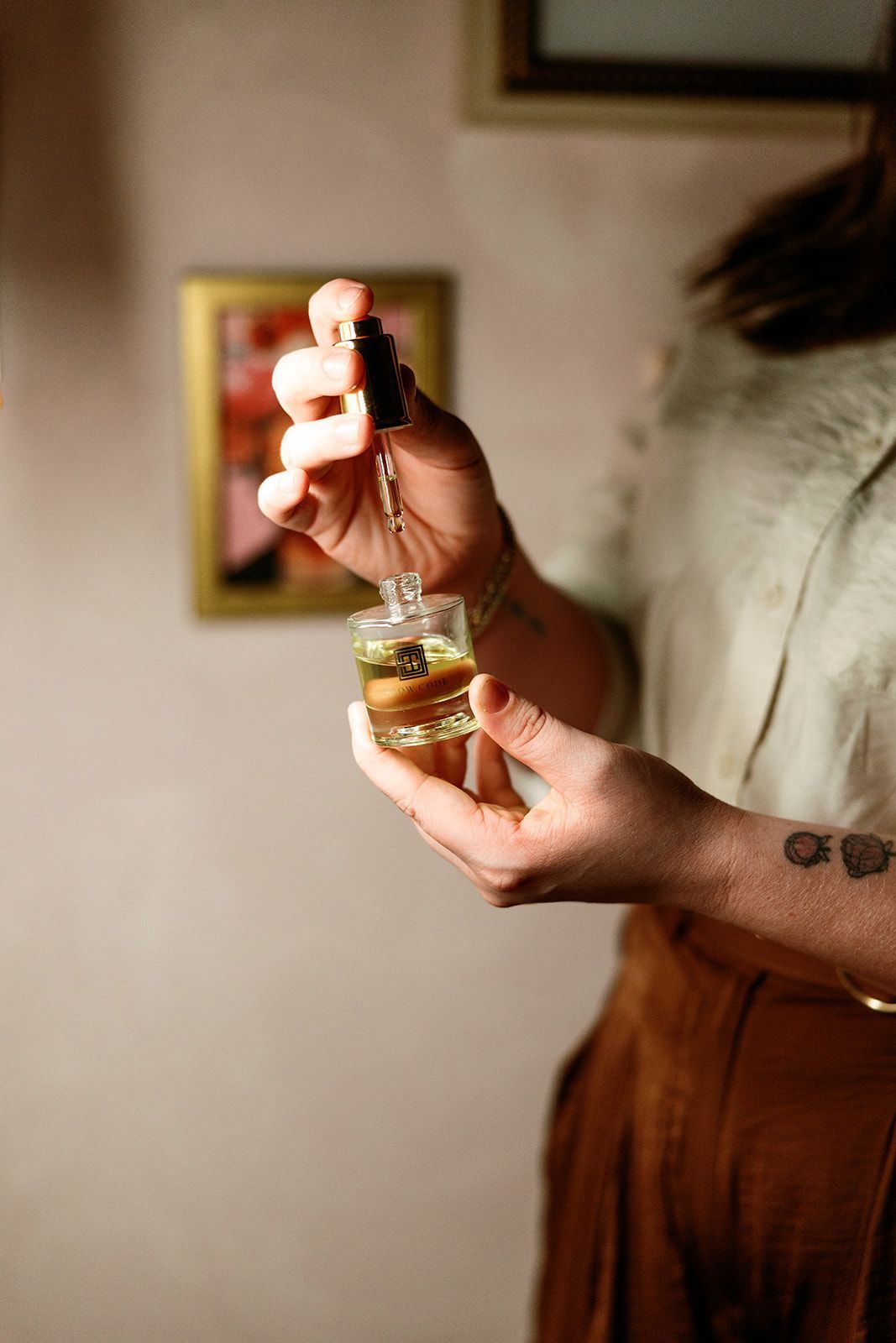 Hands holding a small glass bottle with dropper of liquid, indoors near a blurred framed picture.