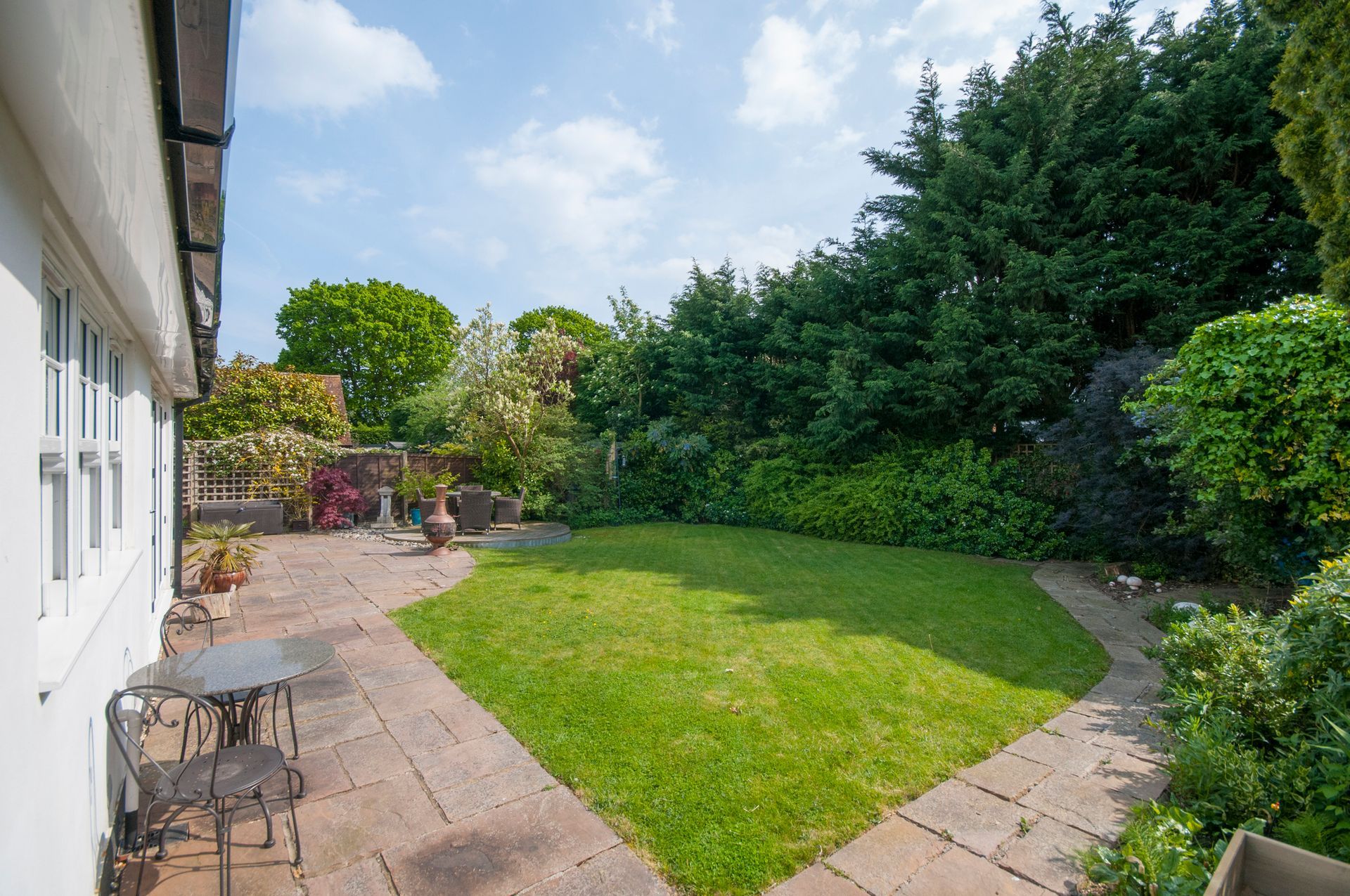 Lawn and patio in a backyard with lush green trees and a white house. Sunny day. Lawn and patio in a backyard with lush green trees and a white house. Sunny day.