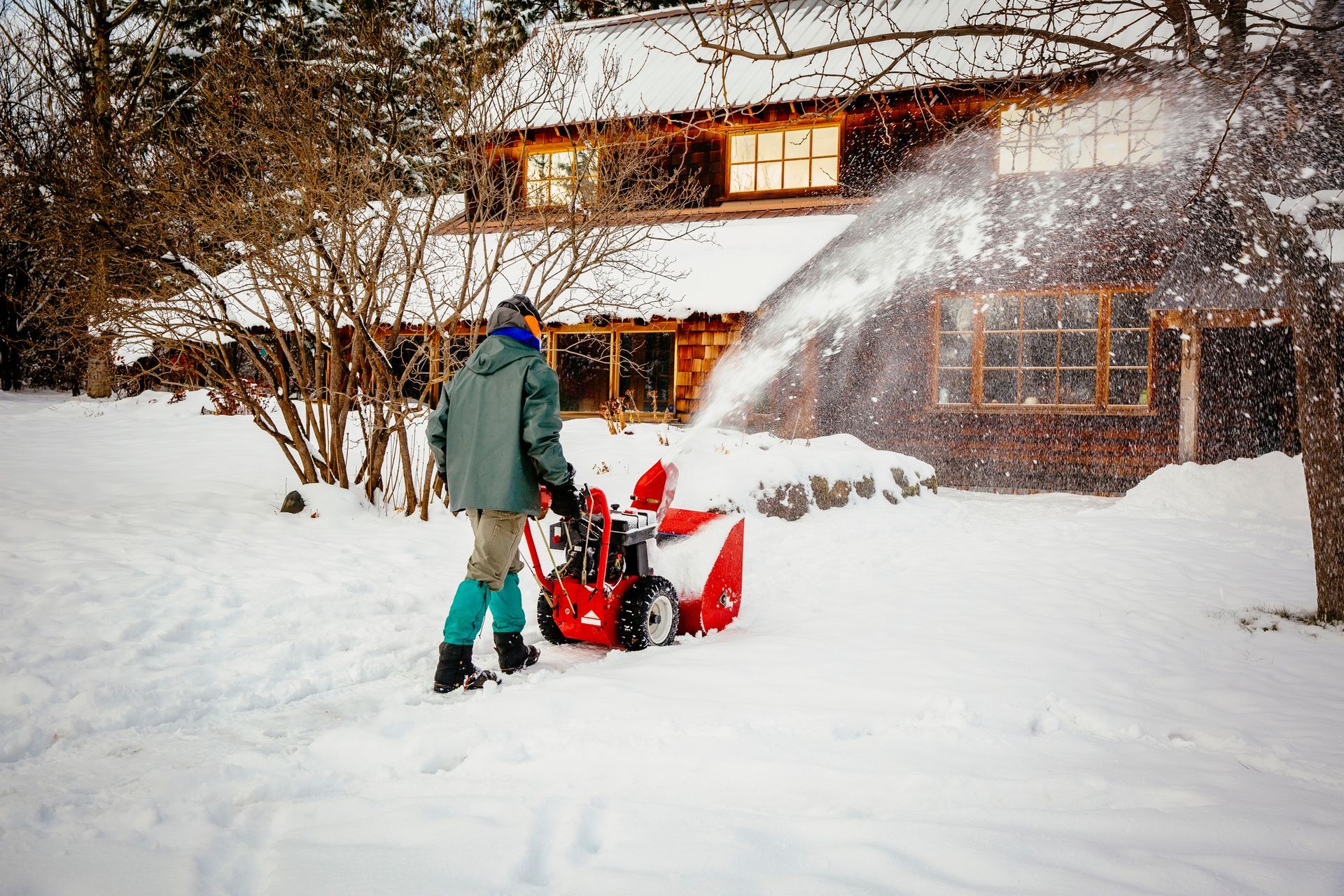 Person using a red snowblower clears snow near a brown house, winter setting. Person using a red snowblower clears snow near a brown house, winter setting.