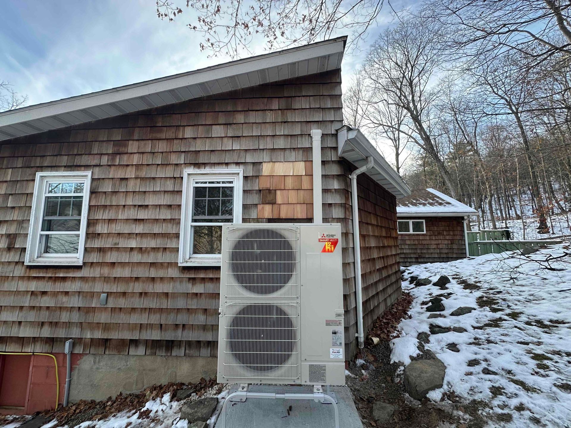 House with an outdoor HVAC unit beside a shingled wall on a snowy hillside