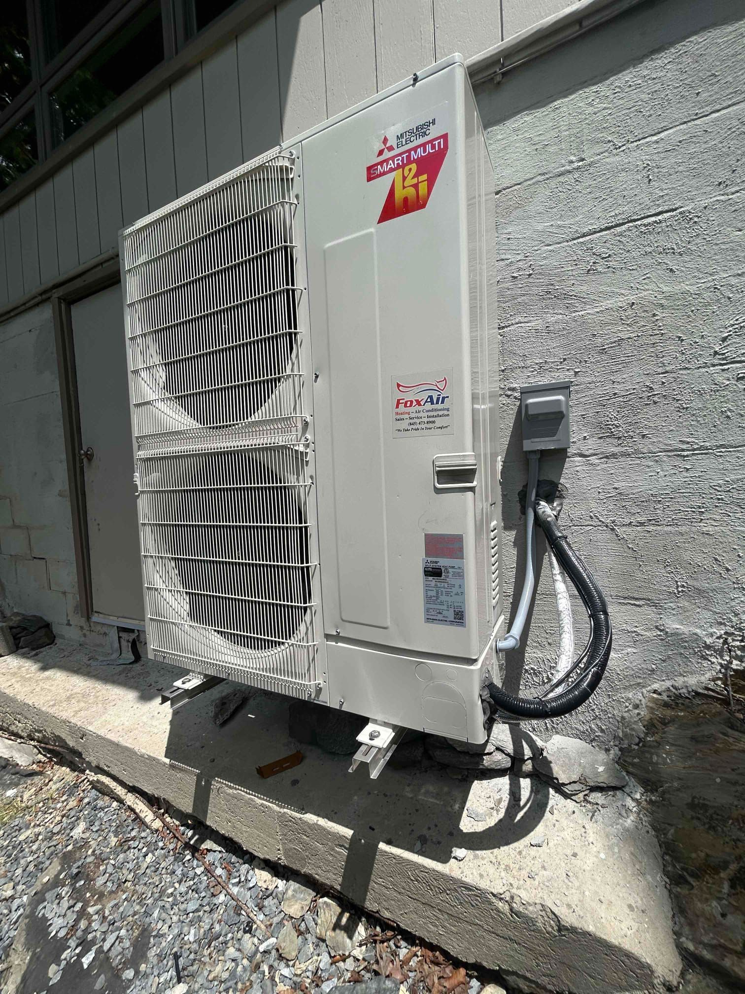 Outdoor wall-mounted air conditioner condenser unit beside a stucco building, with wiring and hoses attached.