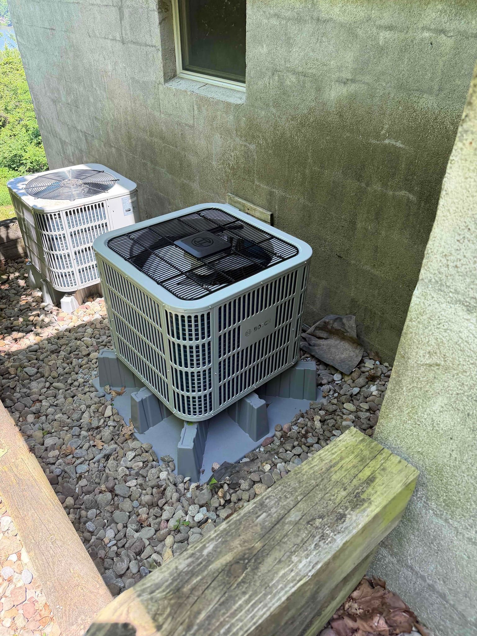 Outdoor HVAC unit with a metal cage beside a building wall and window, on gravel next to a bin