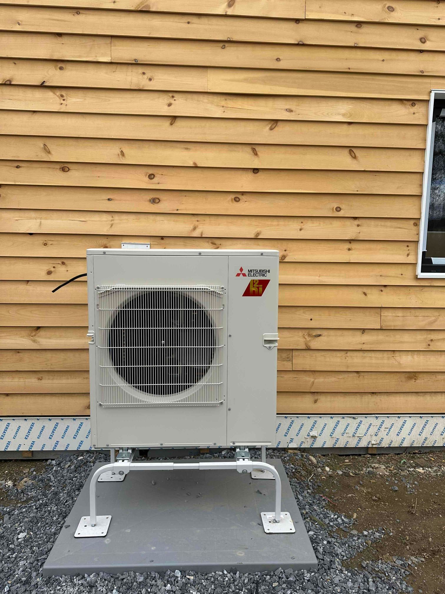 Outdoor HVAC unit on a concrete pad against a tan wood wall.