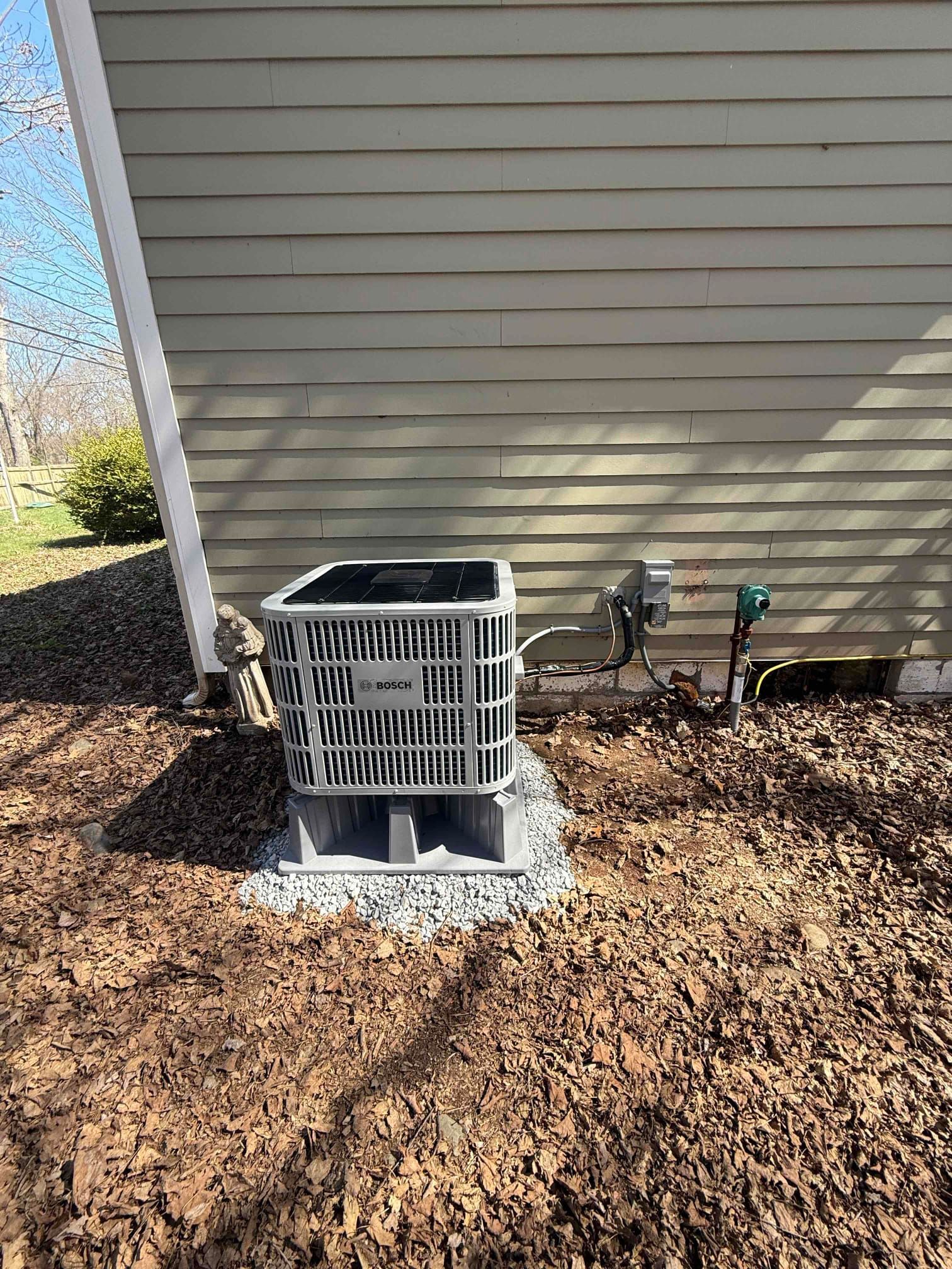 Outdoor air conditioner unit beside a house exterior, with tan siding and mulch around the base.