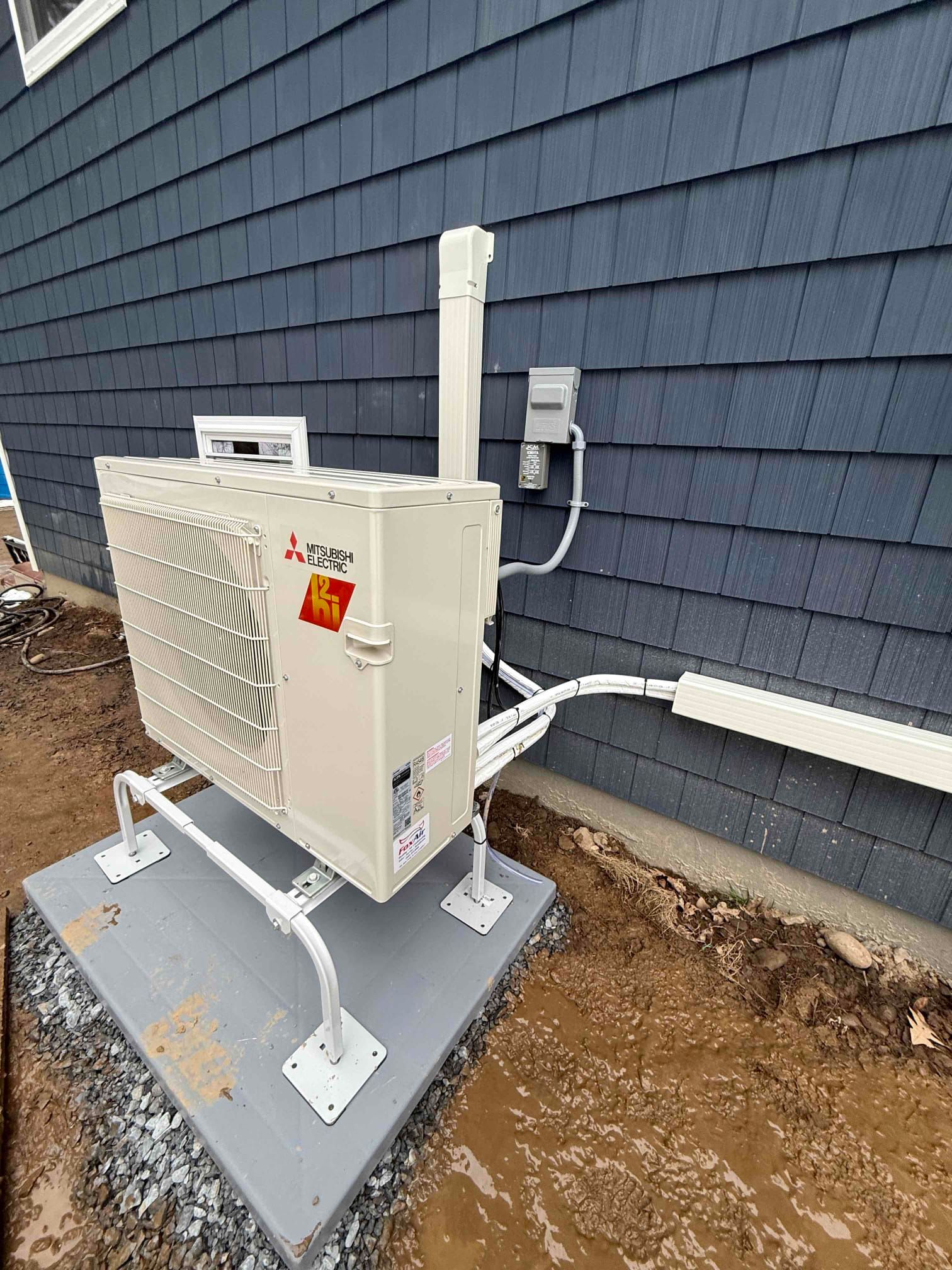 Outdoor HVAC unit on a concrete pad beside a blue house exterior, with conduit and wiring attached.