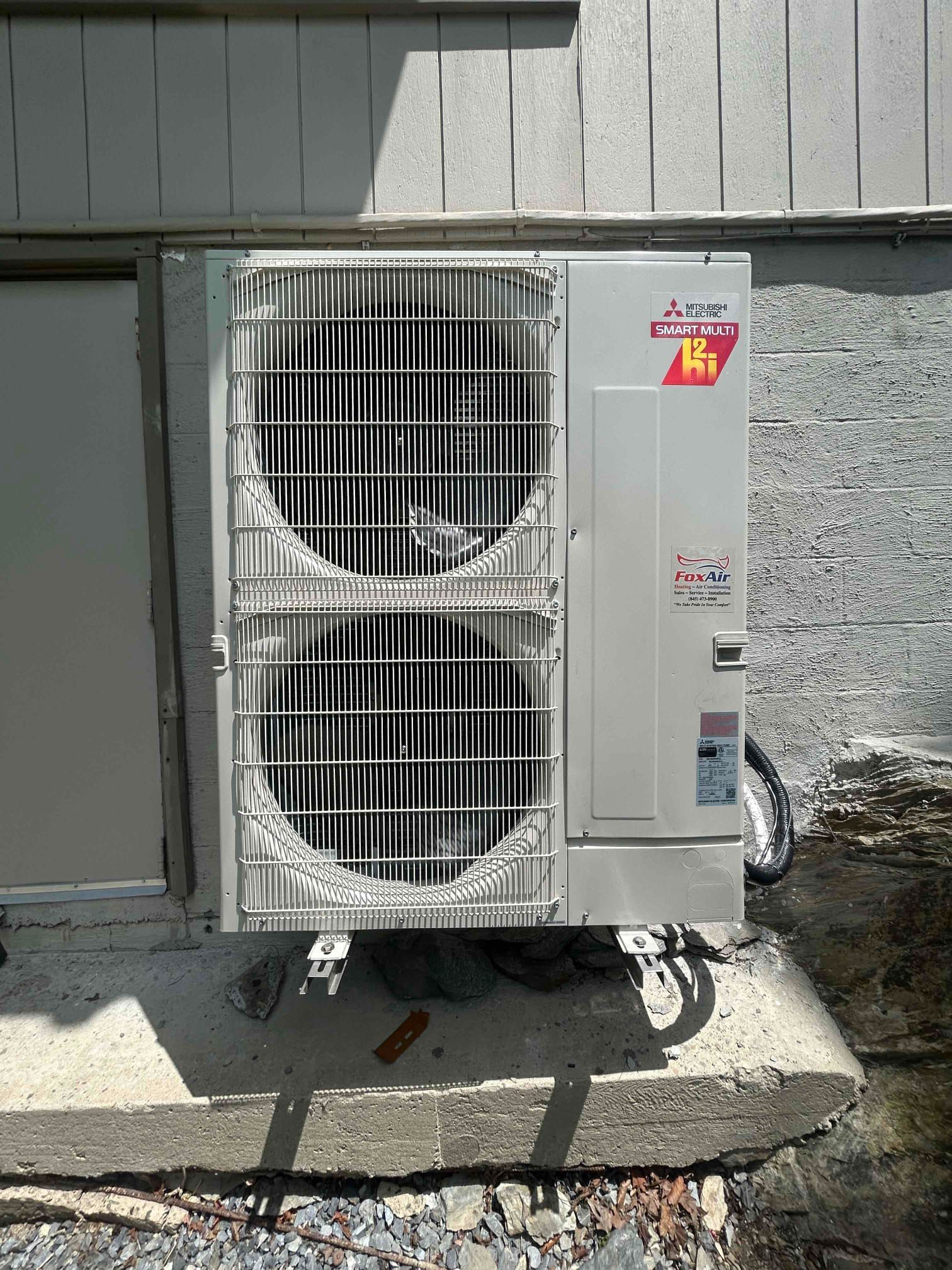 Outdoor air conditioning unit mounted beside a building wall, with a white condenser and metal cage.