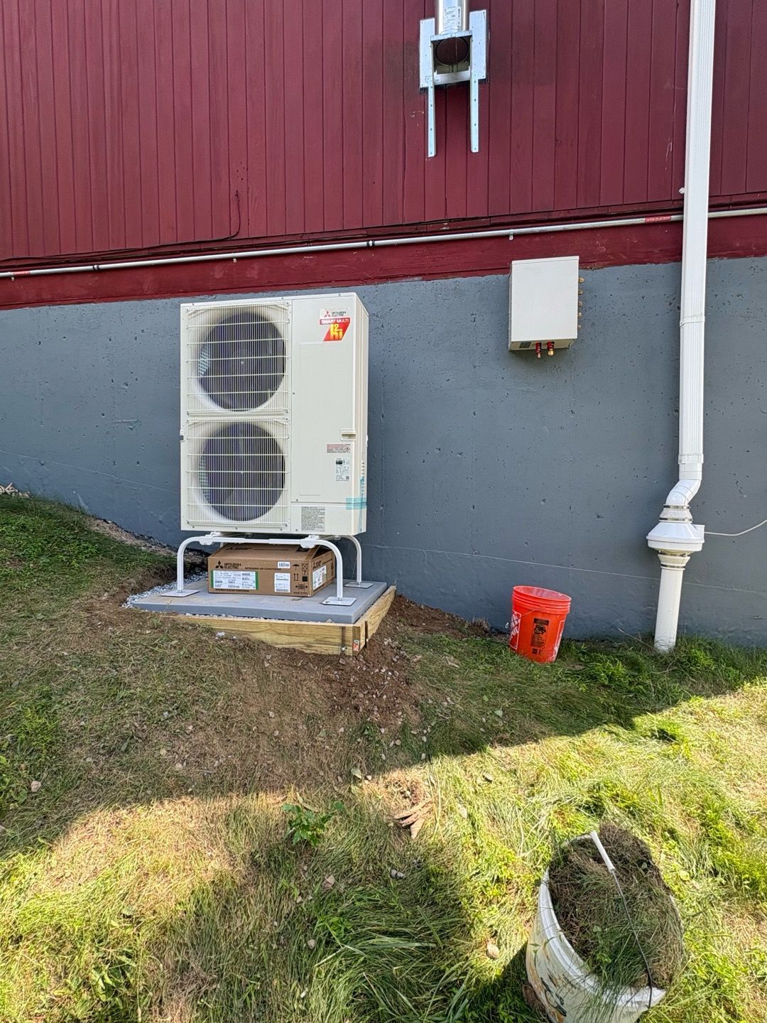 Outdoor heat pump unit beside a red and gray building, with a downspout and small lawn slope.