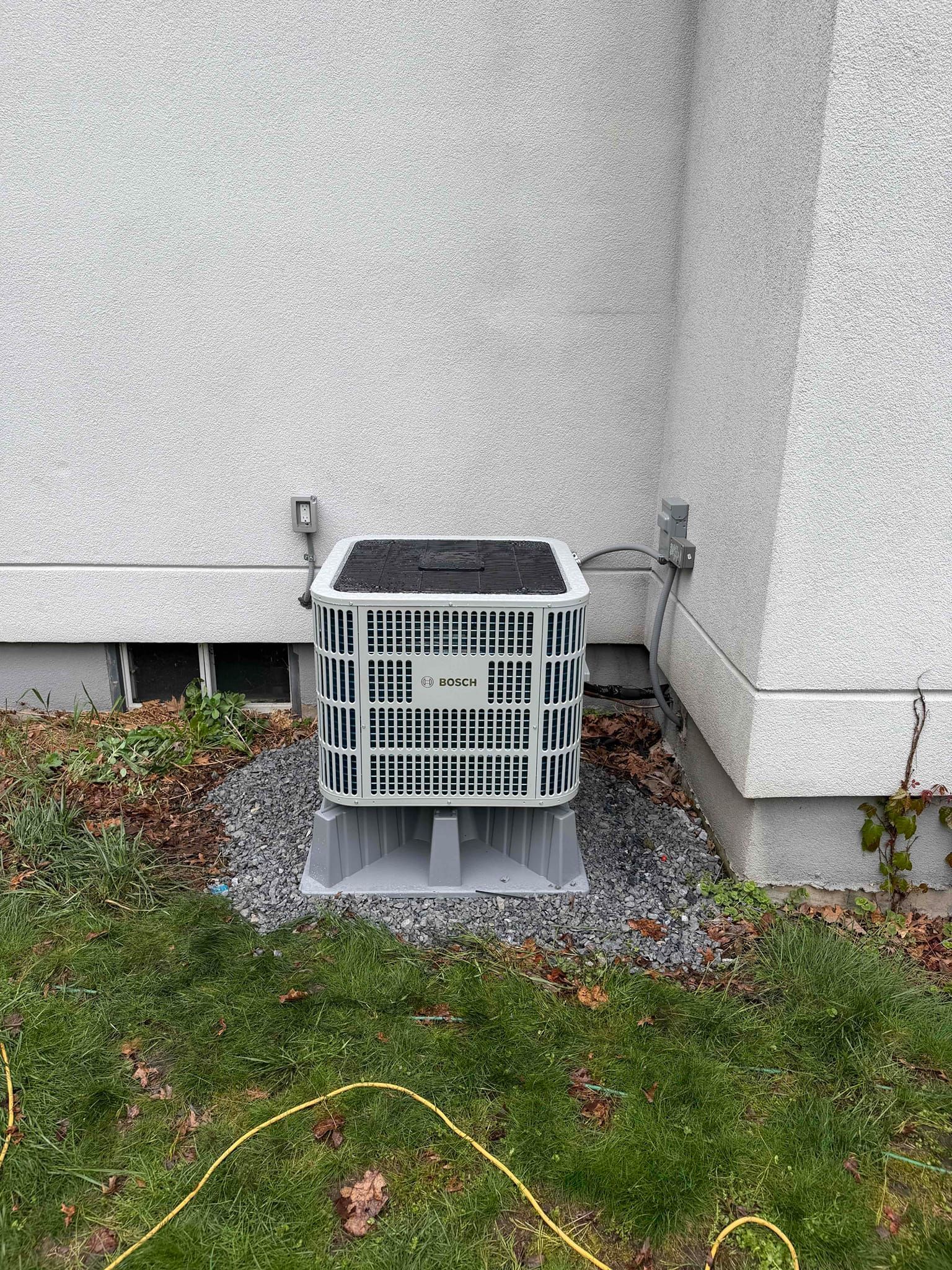 Outdoor HVAC unit beside a house wall, with a white cage and black top on gravel near grass.