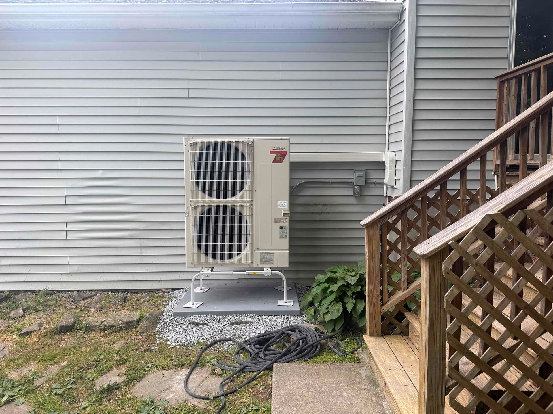 Outdoor HVAC unit beside a house, near white siding, lattice stairs, and a coiled hose on the ground