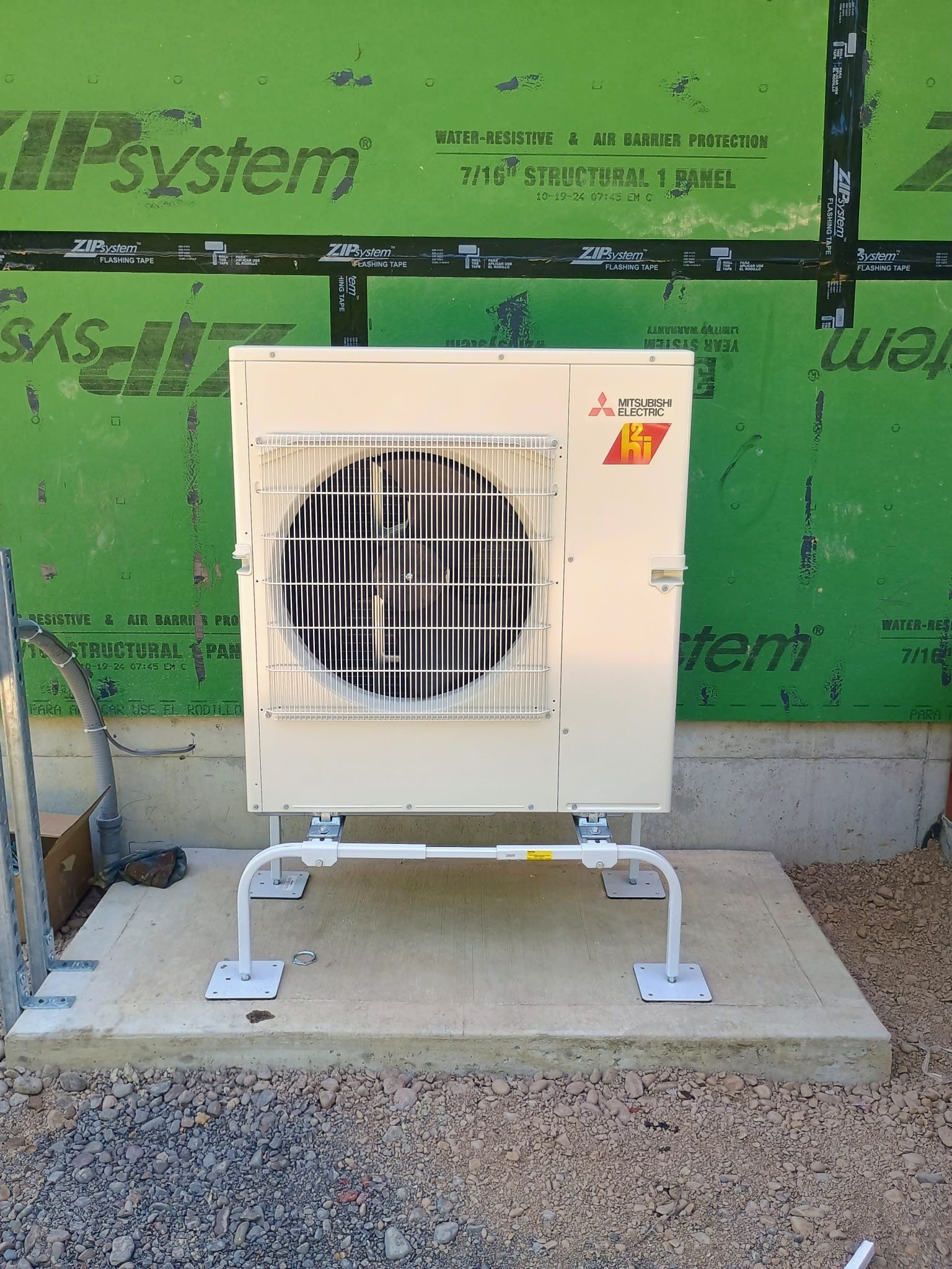 White outdoor HVAC unit on a concrete pad against a green wall with protective bars over the fan opening