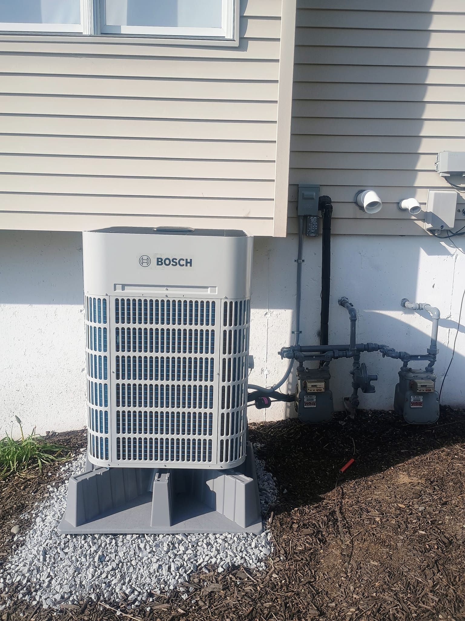 Outdoor Bosch HVAC unit beside a beige house with exposed pipes and gravel ground