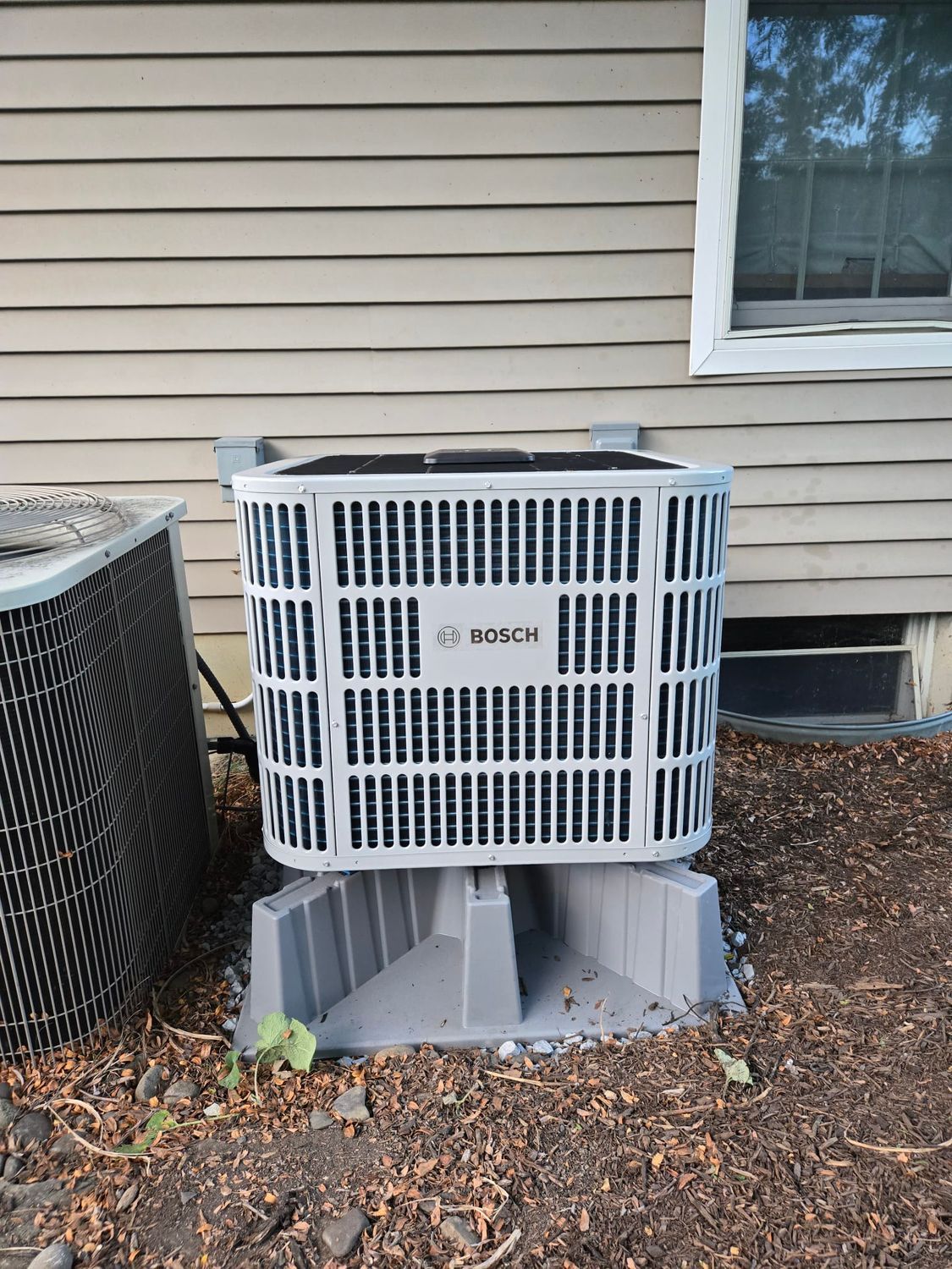 White outdoor air conditioner unit beside a house with beige siding and a window, on a gray platform.