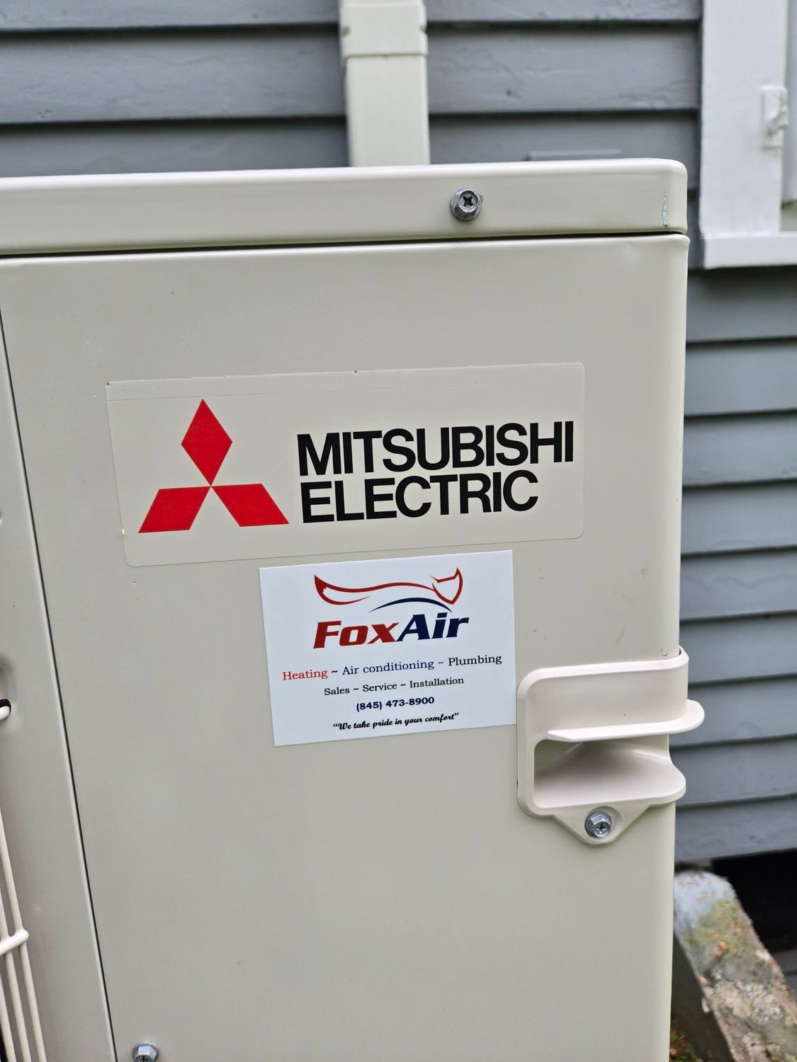 Mitsubishi Electric outdoor HVAC unit on a gray house exterior with a FoxAir service sticker.