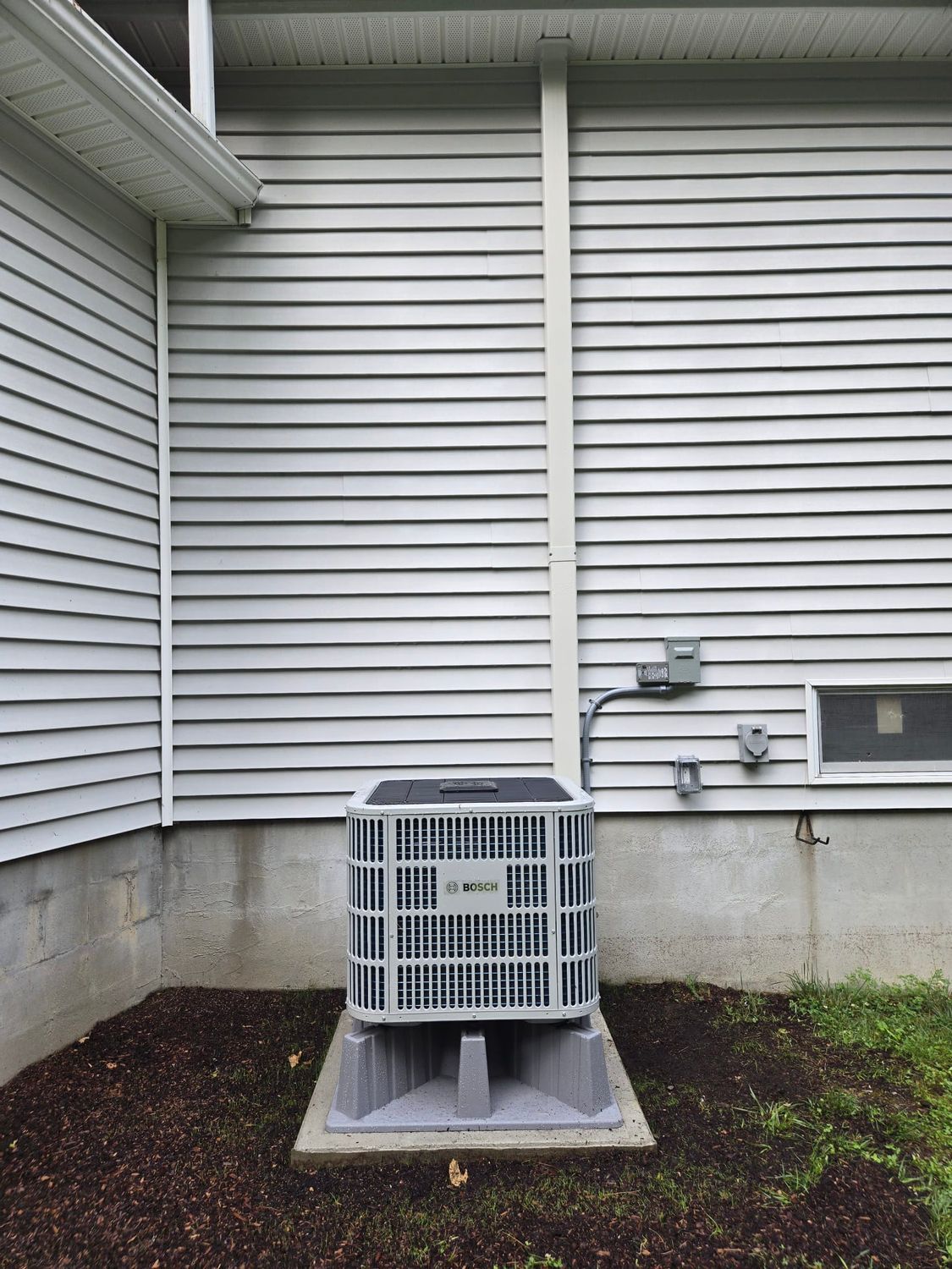 Outdoor HVAC unit beside a house wall, set on a concrete pad under vinyl siding.