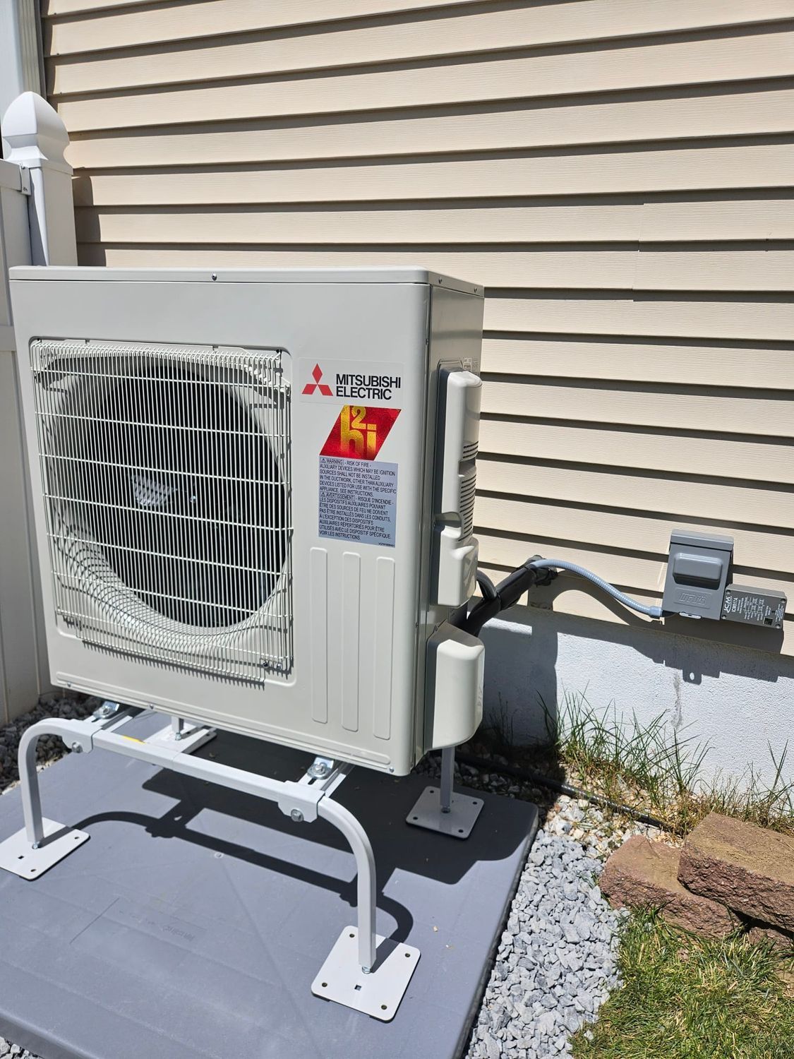 Outdoor air conditioning unit mounted beside a house, with white housing and visible warning labels.