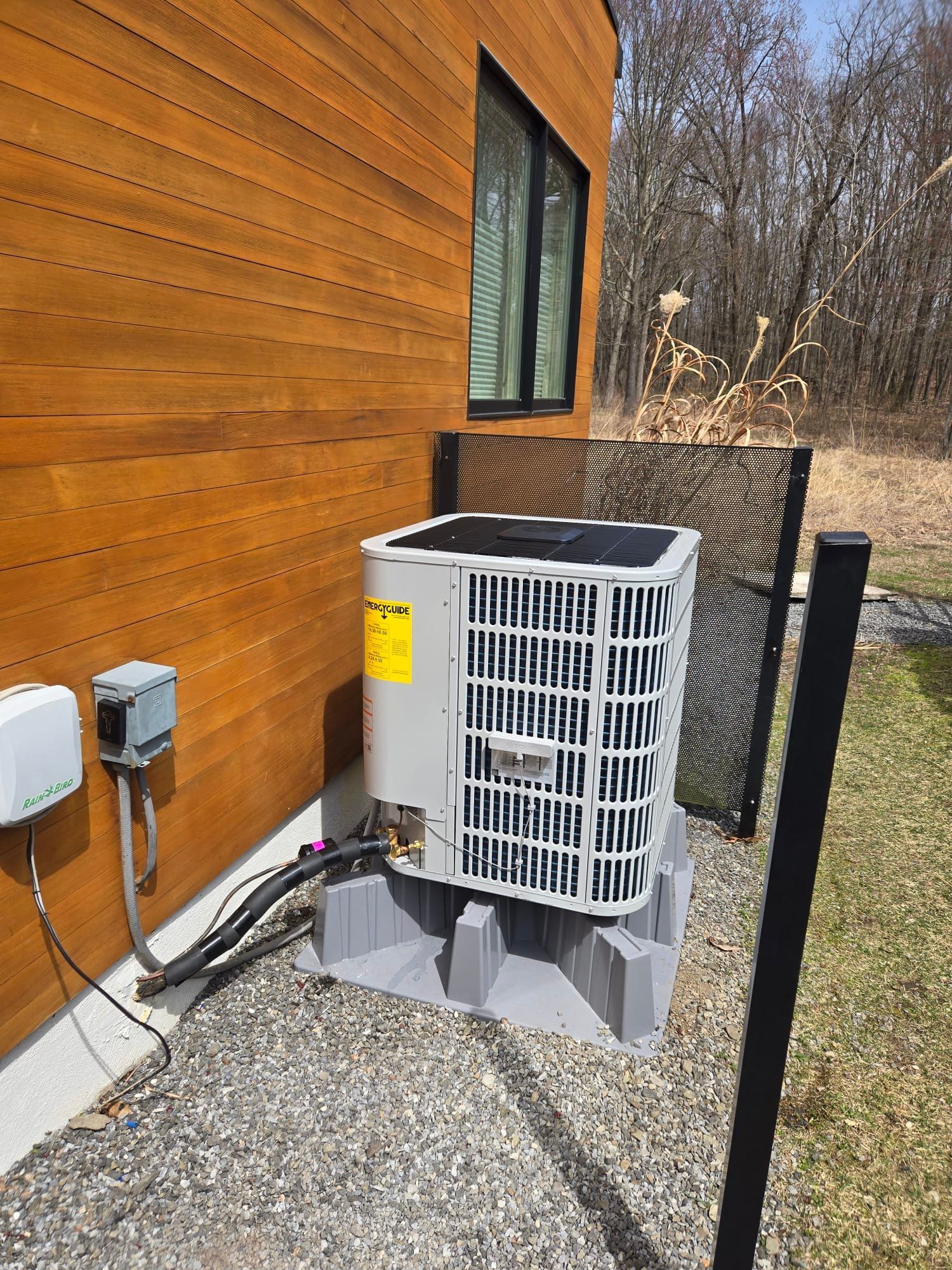Outdoor air conditioner unit beside a wooden house wall on gravel, near a window and yard fence.