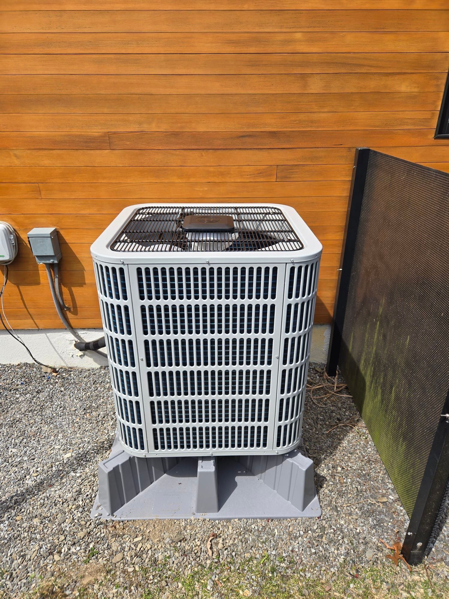 Outdoor air conditioner unit beside an orange wooden wall and a black privacy screen on gravel.