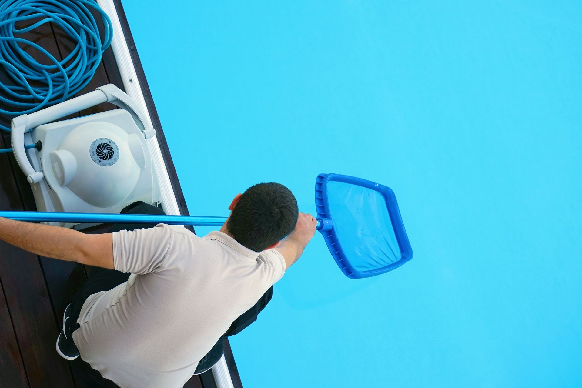 Person using a pool net and cleaning equipment next to a bright blue swimming pool.