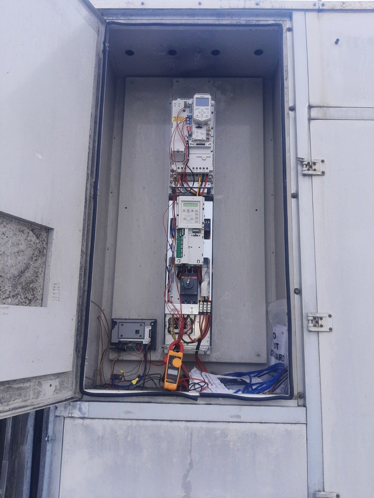 An open utility cabinet contains vertical electrical equipment, including a meter and a multimeter resting at the bottom.