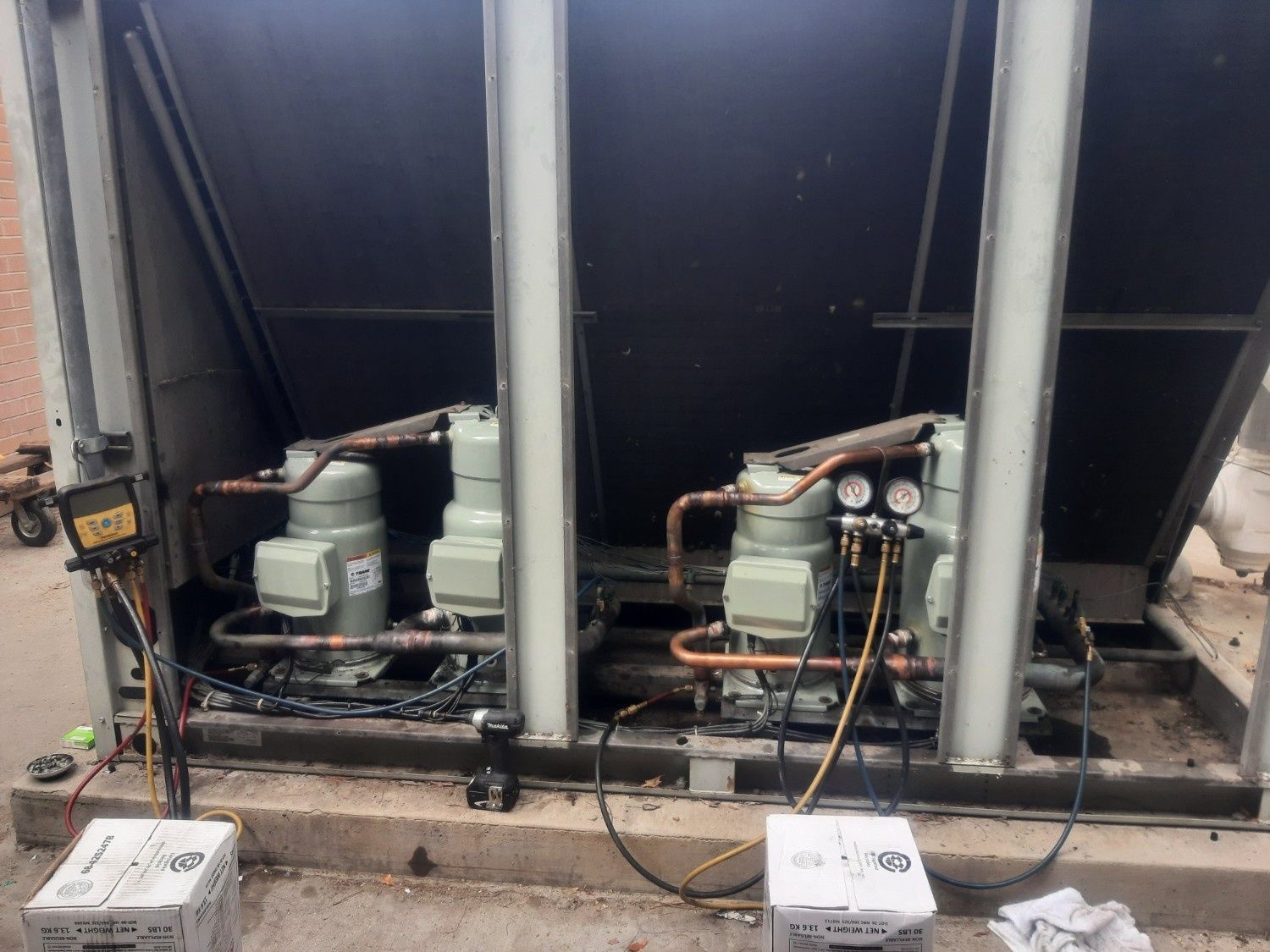 Two HVAC compressors installed outdoors on a concrete base, connected with copper piping and diagnostic pressure gauges.