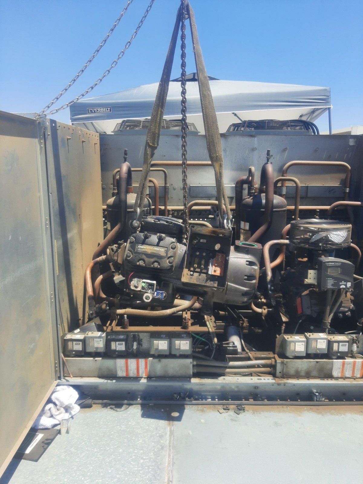 A heavy-duty industrial compressor being lifted by chains from inside a metal cabinet outdoors under a clear blue sky.