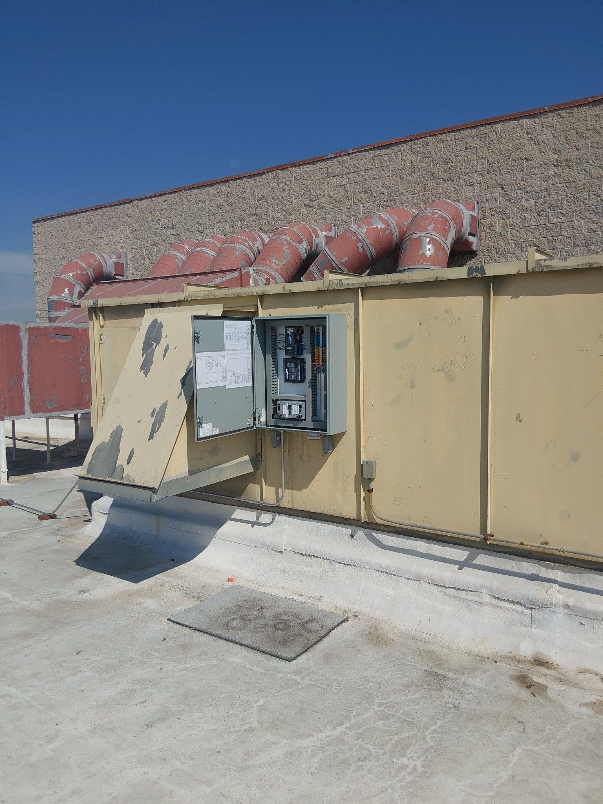 A rooftop HVAC unit with an open electrical control panel box, set against a stone-textured wall under a clear sky.