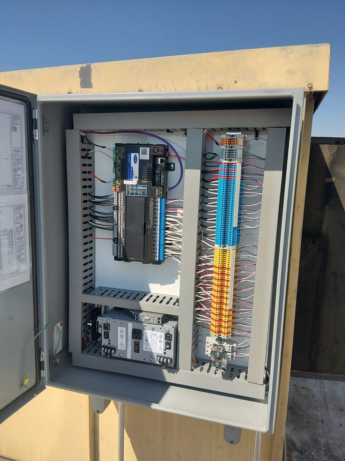 An open electrical control box mounted outdoors, showing a PLC, wiring terminal blocks, and power supply on a white backpanel.