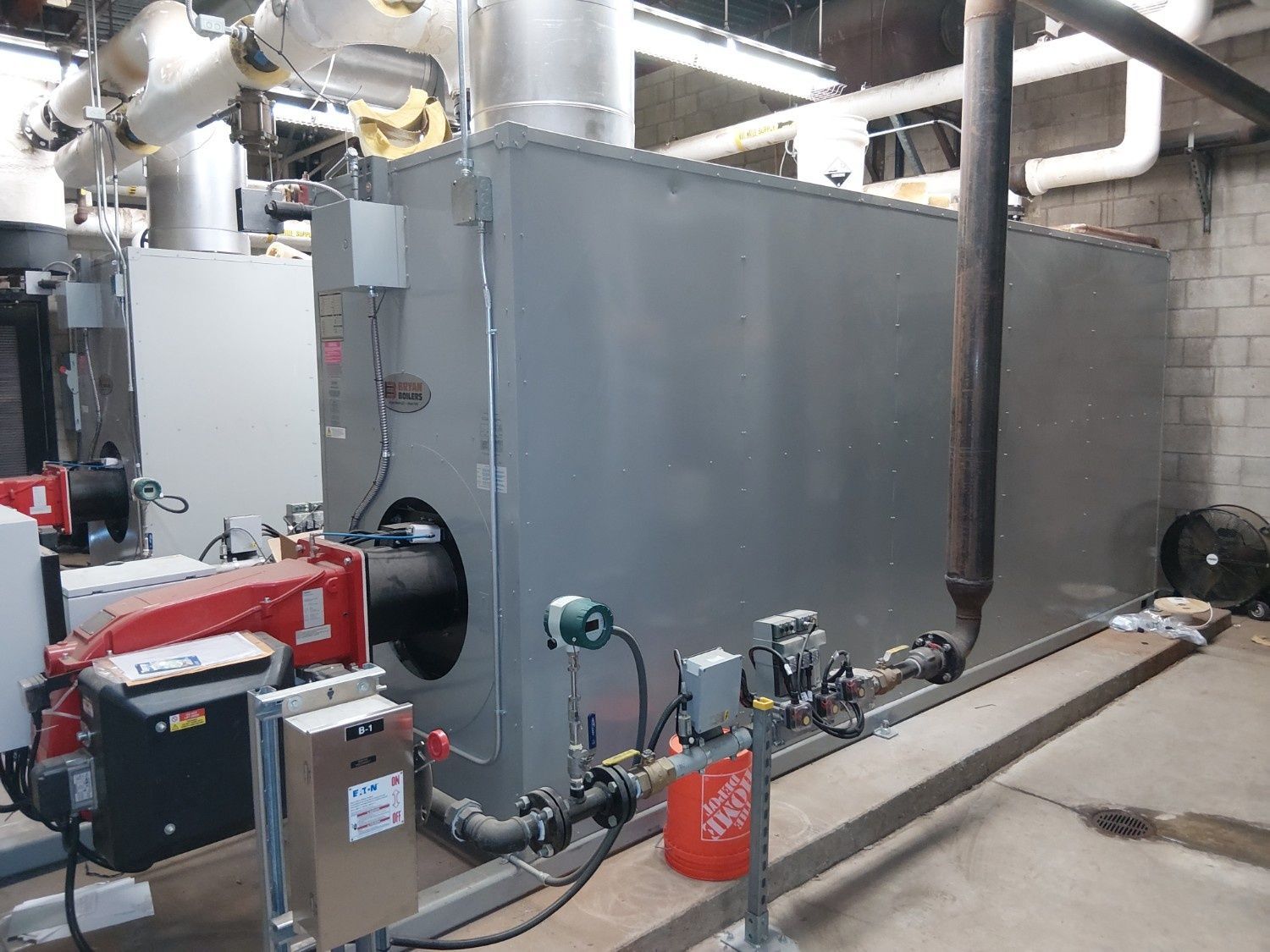 A large gray industrial boiler with a red burner unit, set in a mechanical room with pipes and an orange bucket nearby.