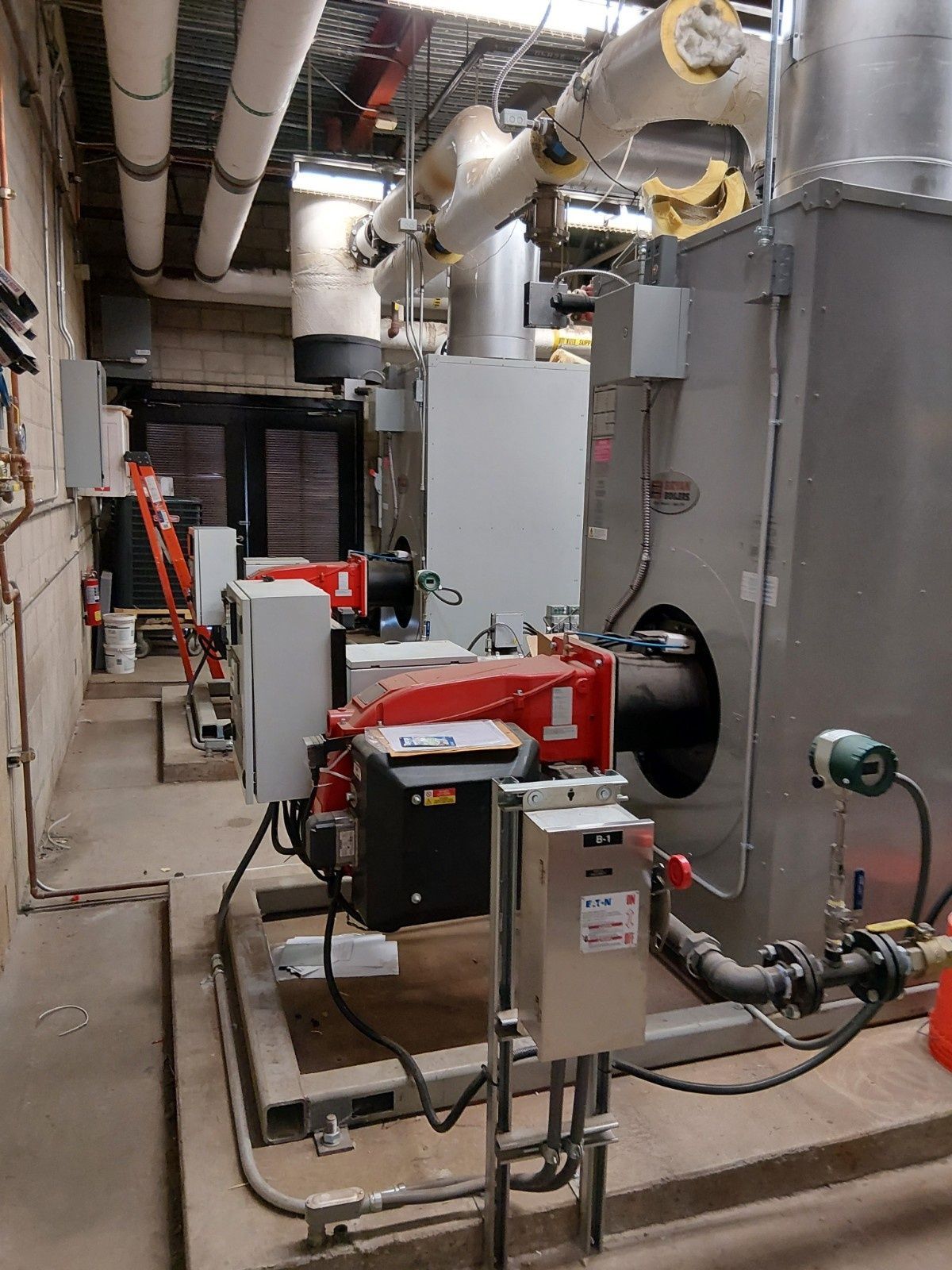 A utility room featuring large industrial boilers with red burners, metal ducting, and piping on a concrete floor.