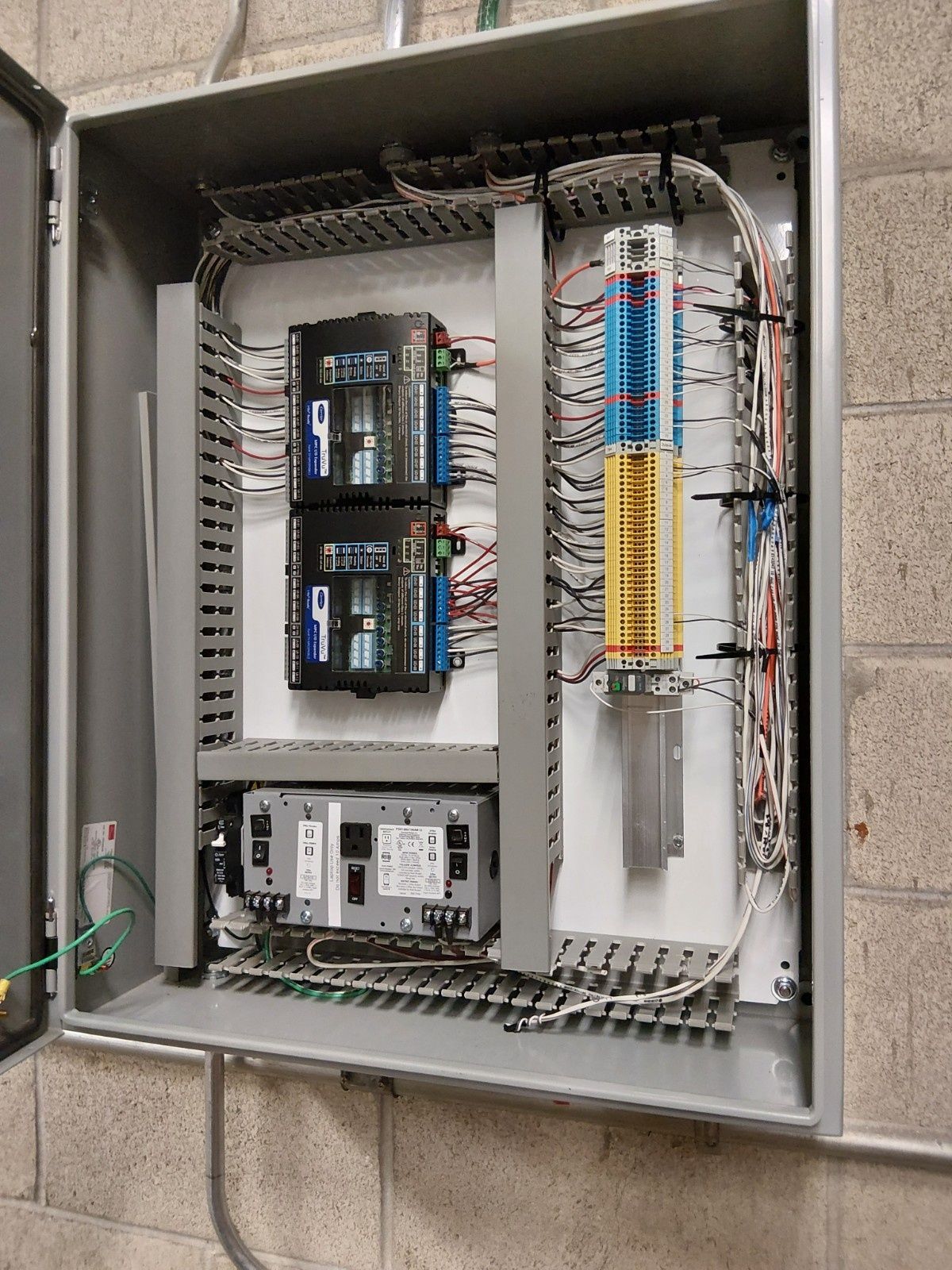 An open electrical control panel mounted on a concrete wall, featuring two black controllers and a terminal block strip.