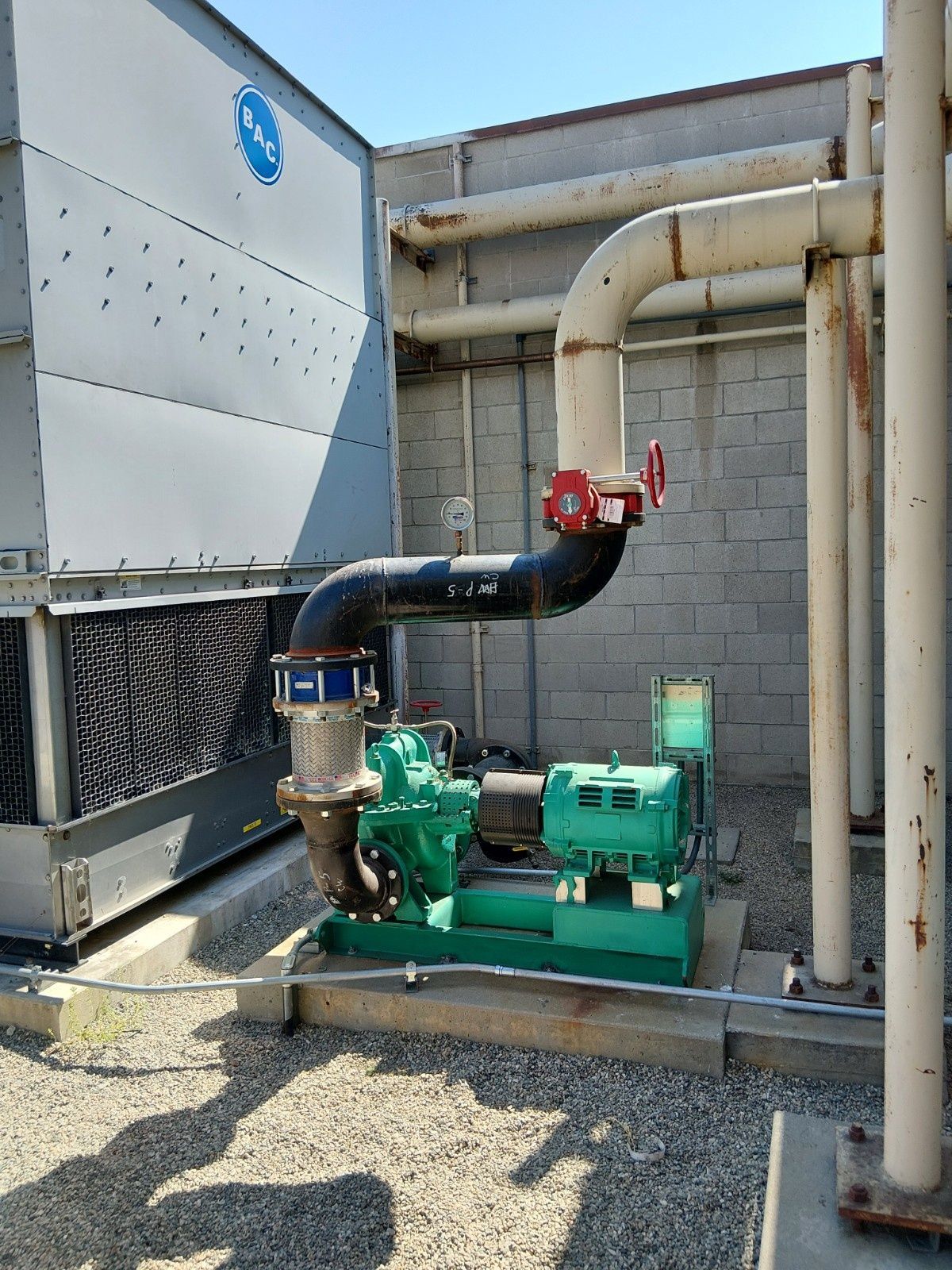 A green industrial pump connected to piping and a cooling tower outdoors on a concrete pad.