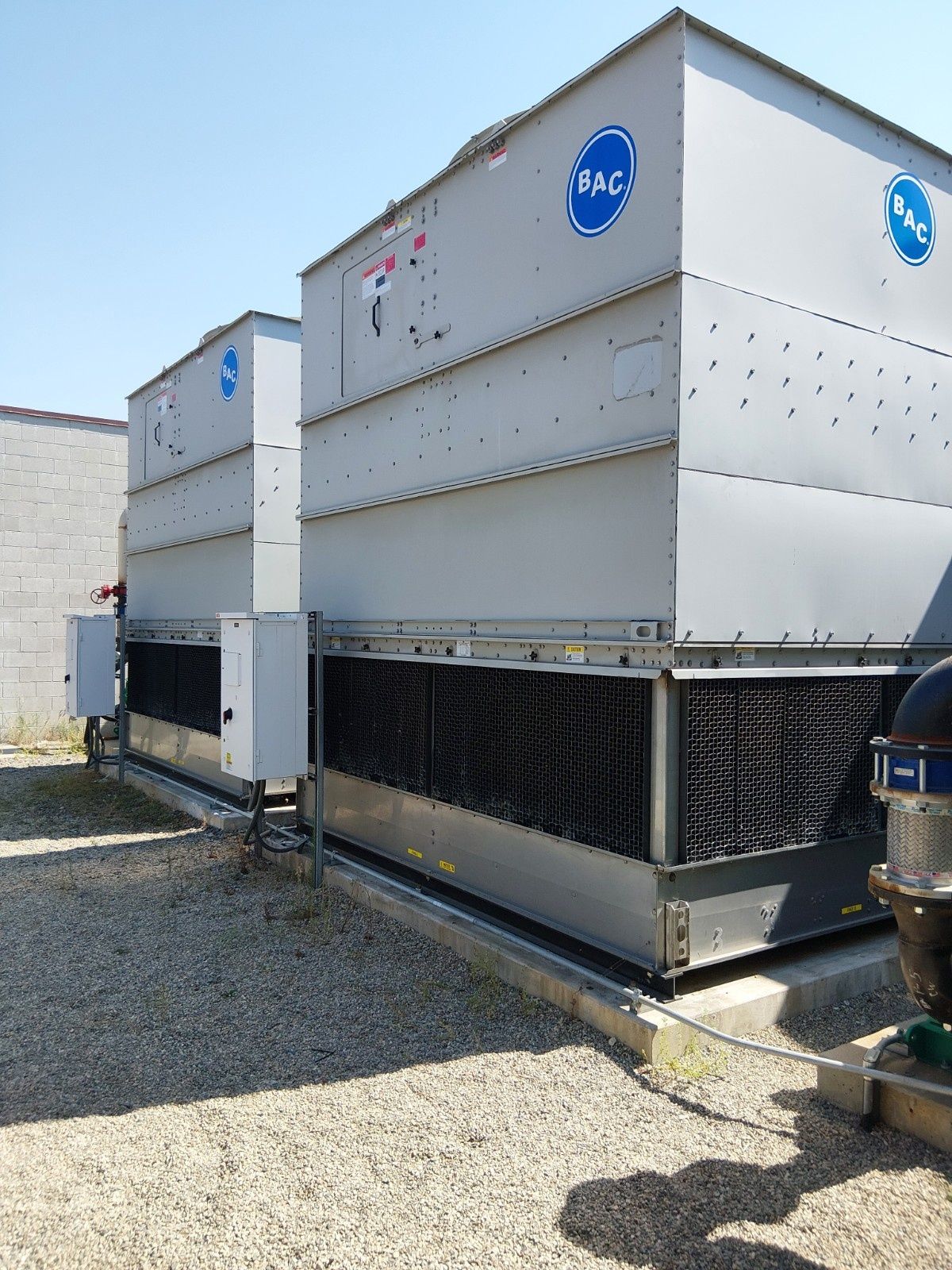 Two large, grey rectangular industrial cooling towers sitting on a gravel lot under a clear blue sky.
