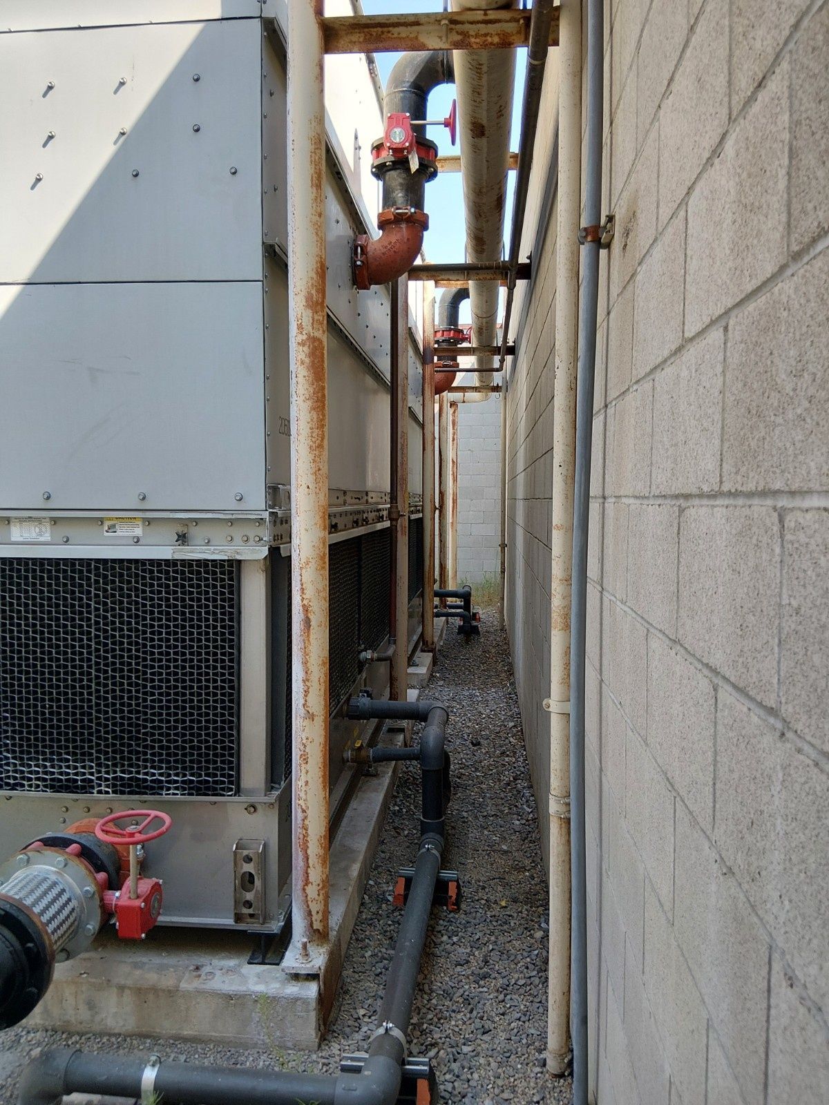 Industrial HVAC cooling tower unit located in a narrow outdoor corridor between a gray metal wall and a cinder block wall.