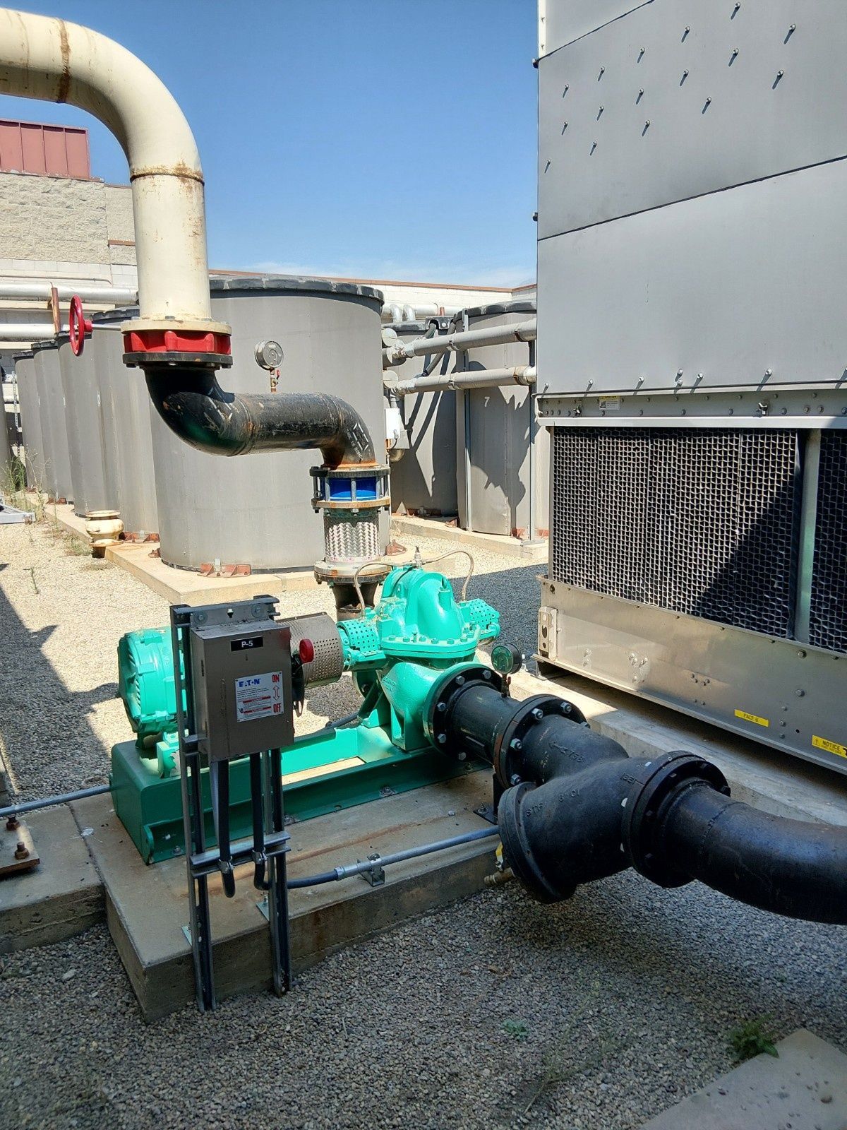 A green industrial water pump sits on a concrete pad next to a cooling tower and grey storage tanks on a gravel surface.