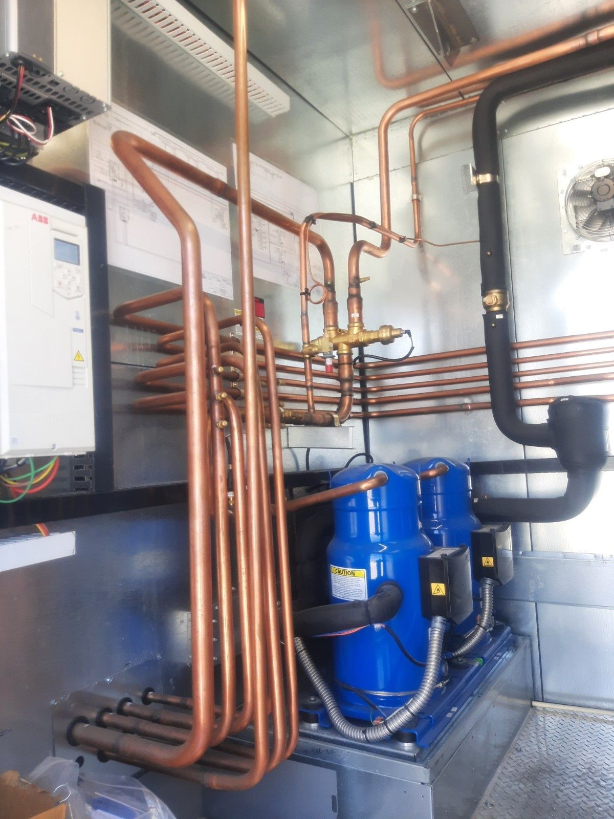 Copper pipes and two blue refrigeration compressors inside a mechanical room with a white control panel on the left.