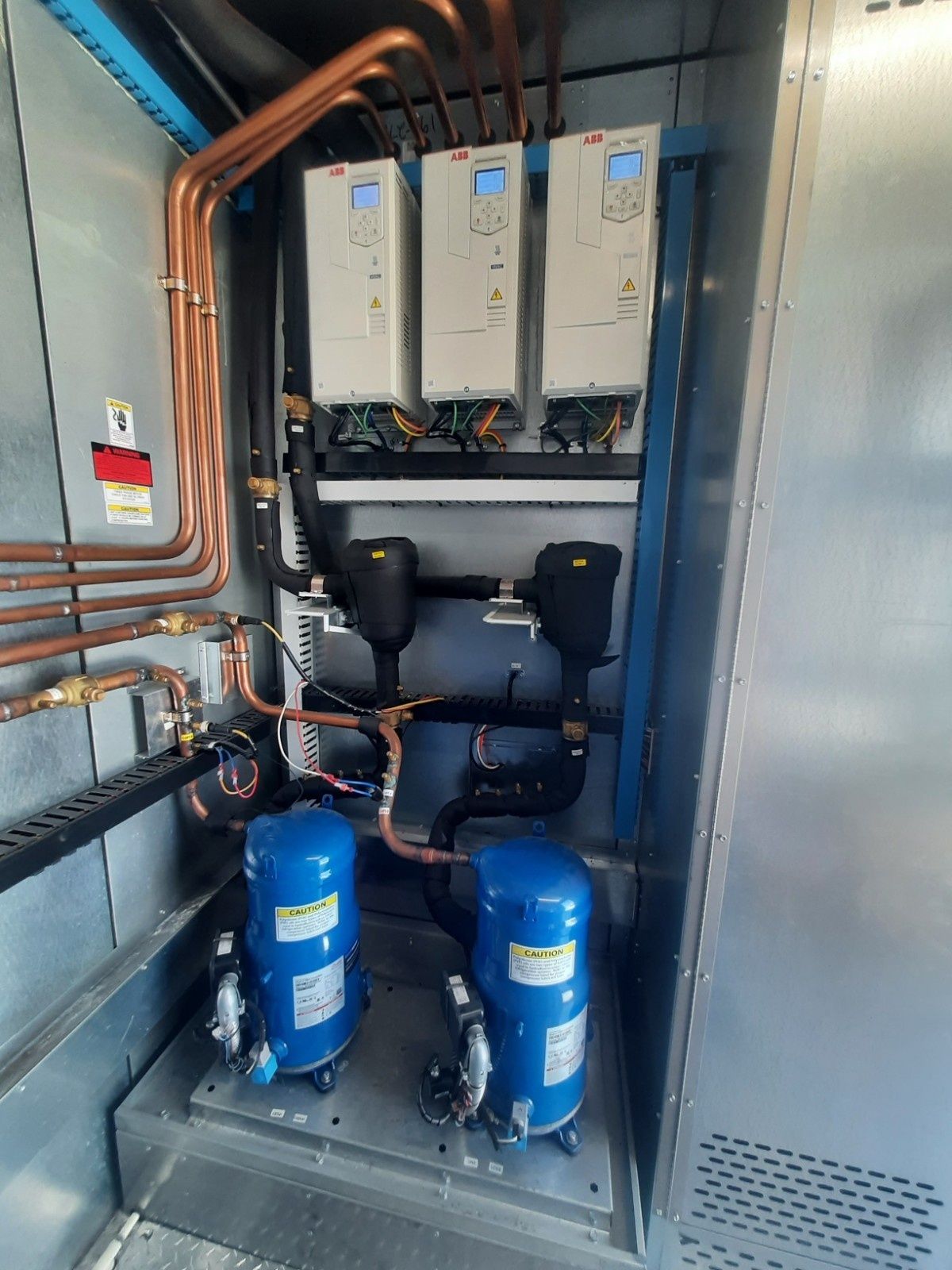 Two blue industrial compressors connected by copper piping below three electronic control units inside a metal cabinet.