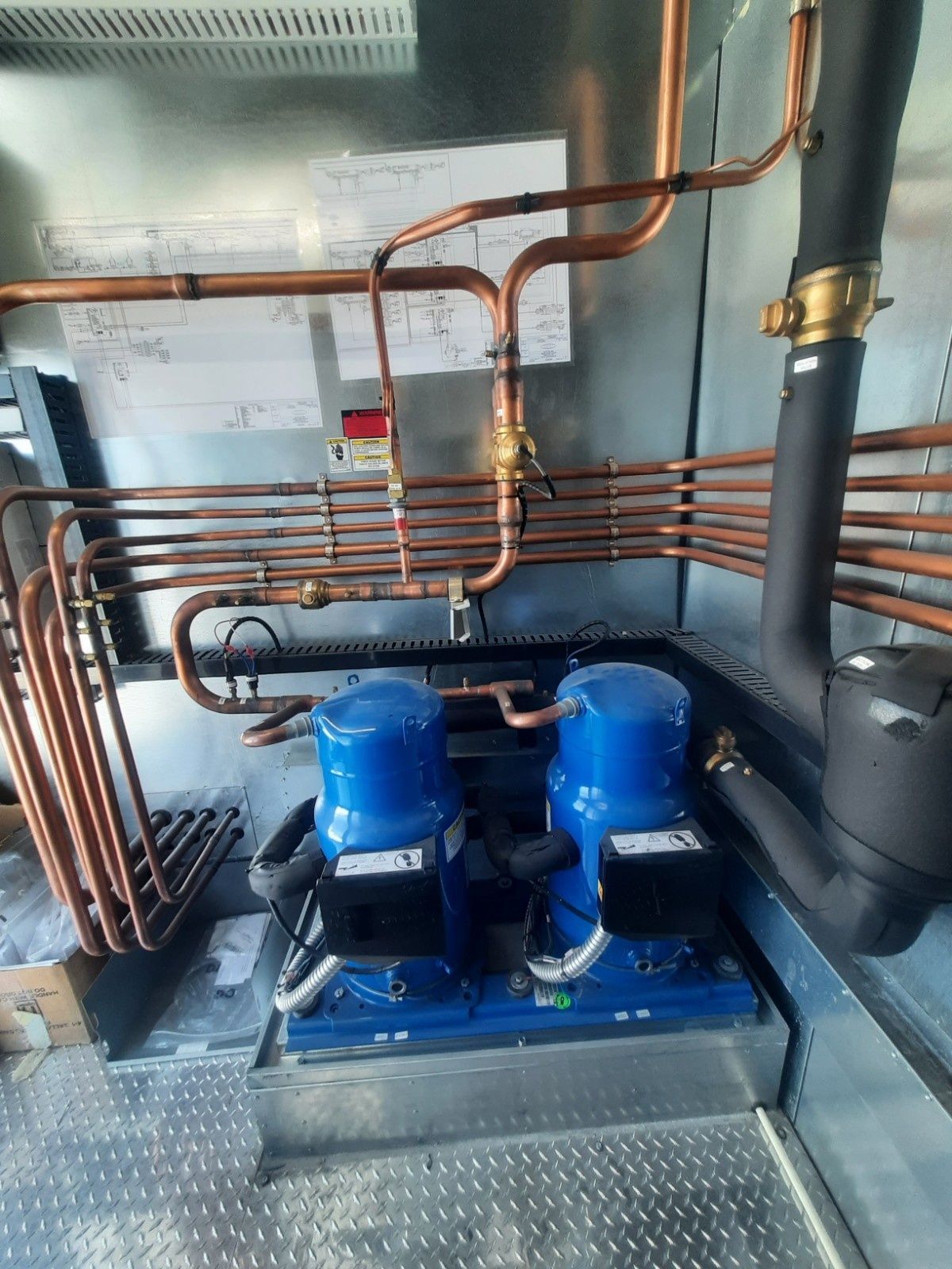 Two blue refrigeration compressors connected to complex copper pipework inside a metal-walled utility room.