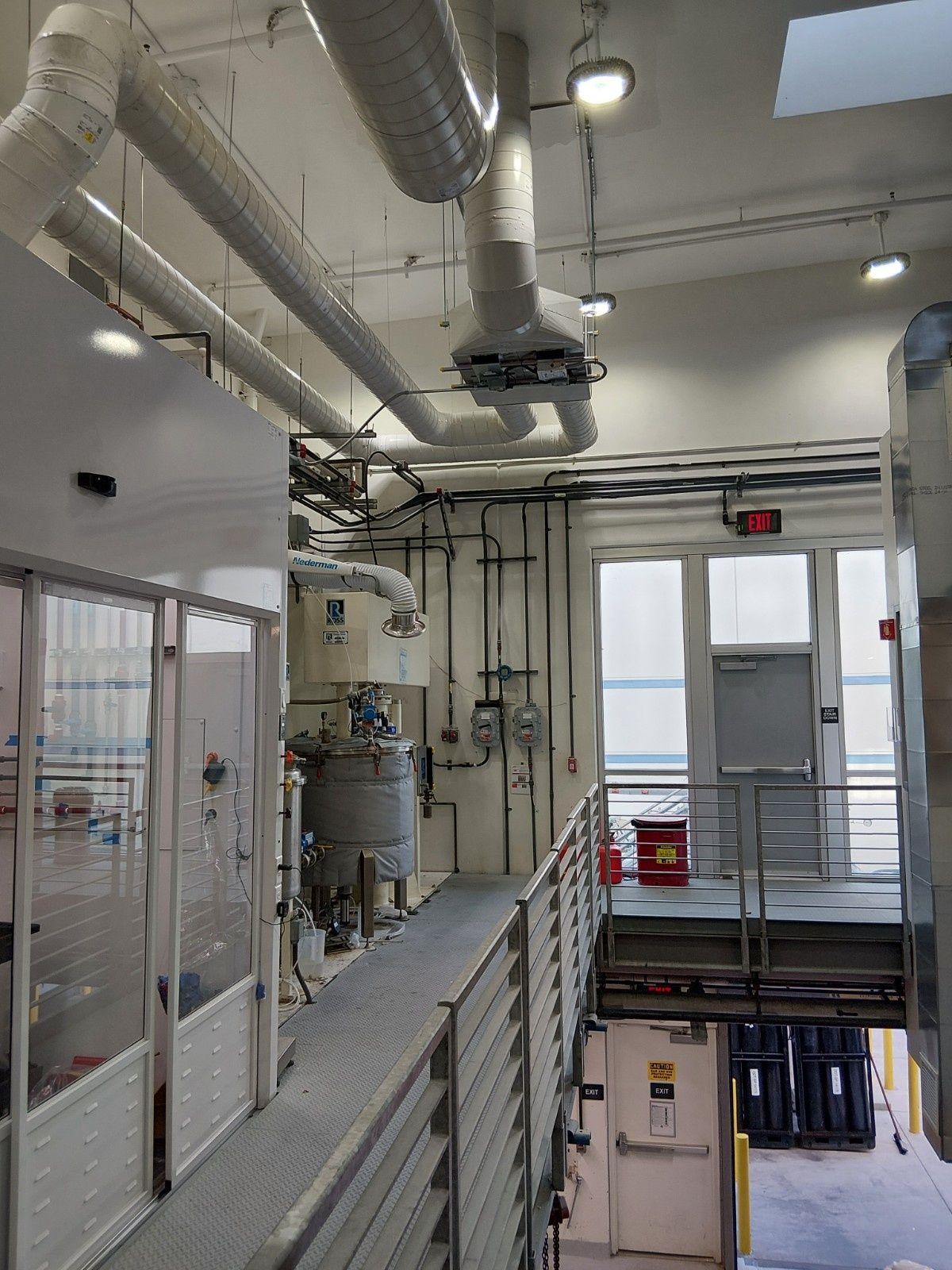 An industrial facility with metal catwalks, large overhead ventilation ducts, machinery, and doors under bright lighting.