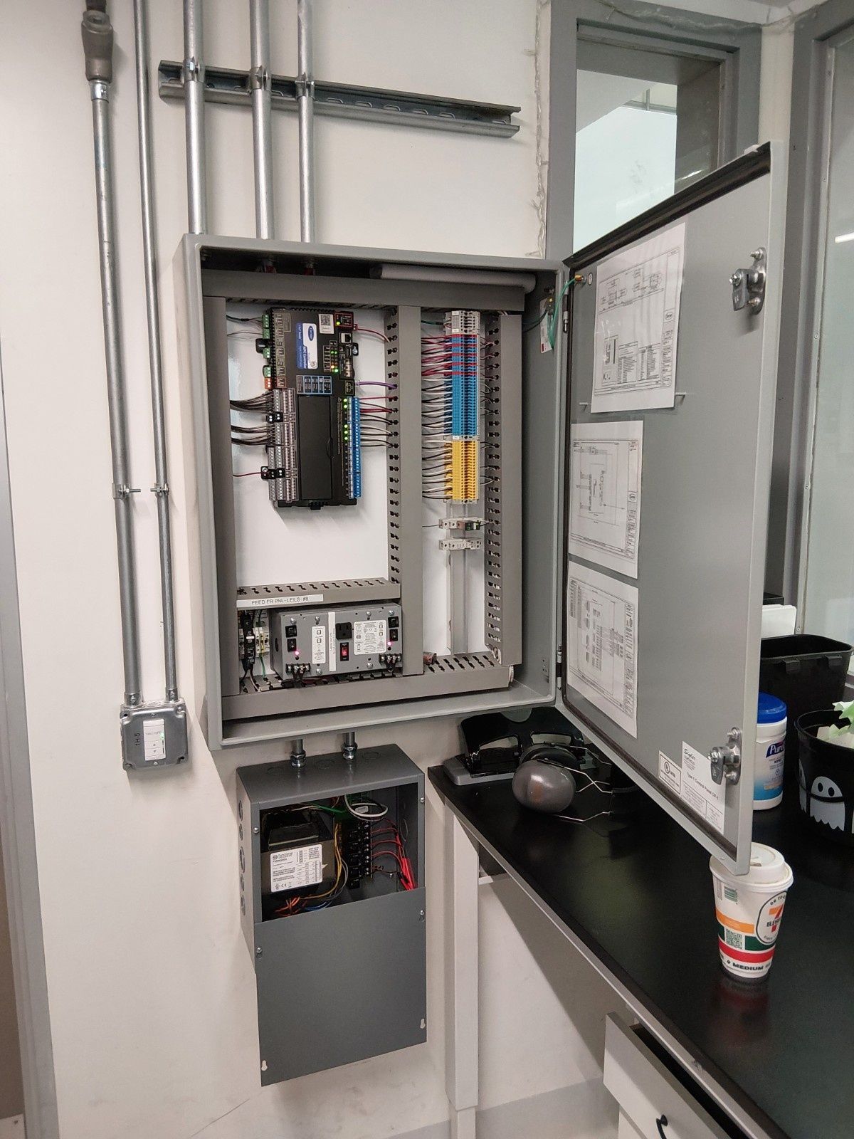 A gray electrical control panel with its door open, mounted on a white wall above a smaller enclosure and a black counter.