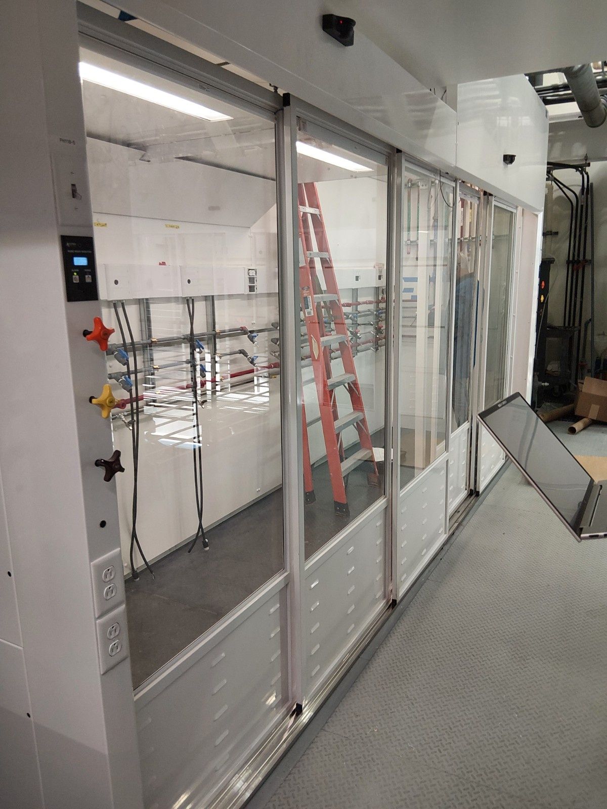 A modular cleanroom or lab enclosure with sliding glass panels, an internal ladder, and wall-mounted electrical outlets.