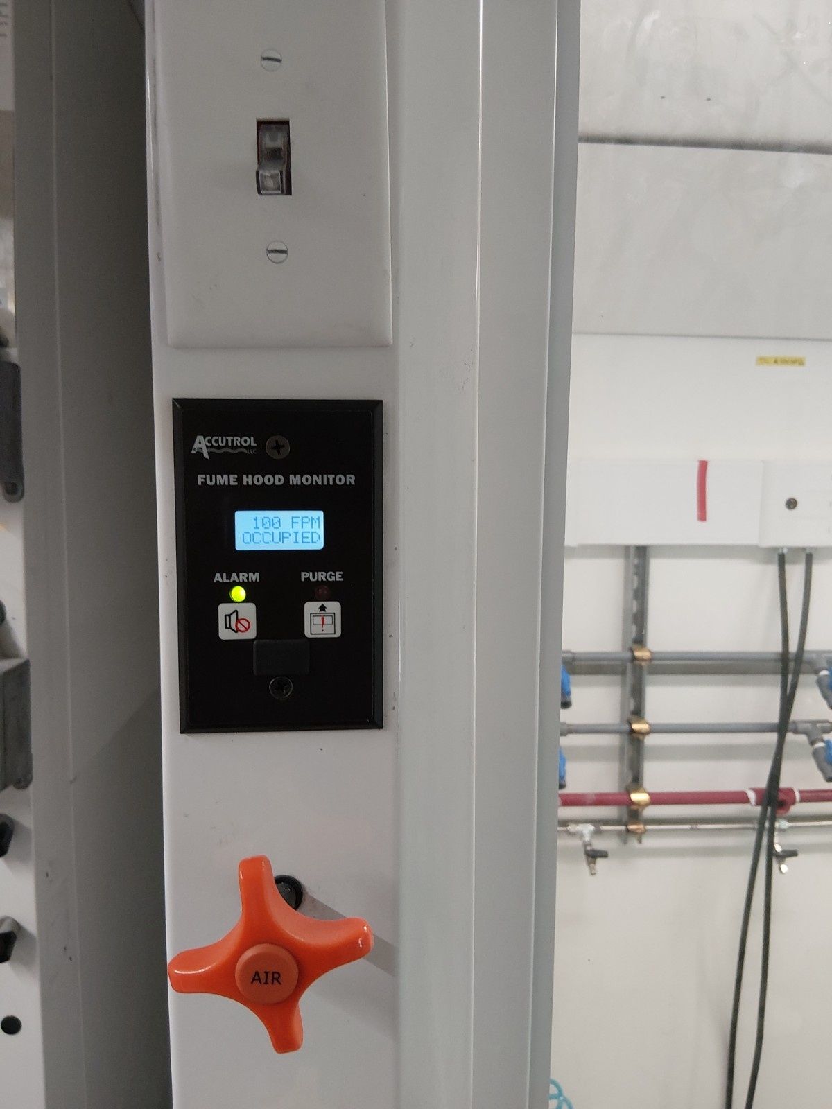 A fume hood monitor display with a green status light, mounted on a white wall above a large orange manual control knob.